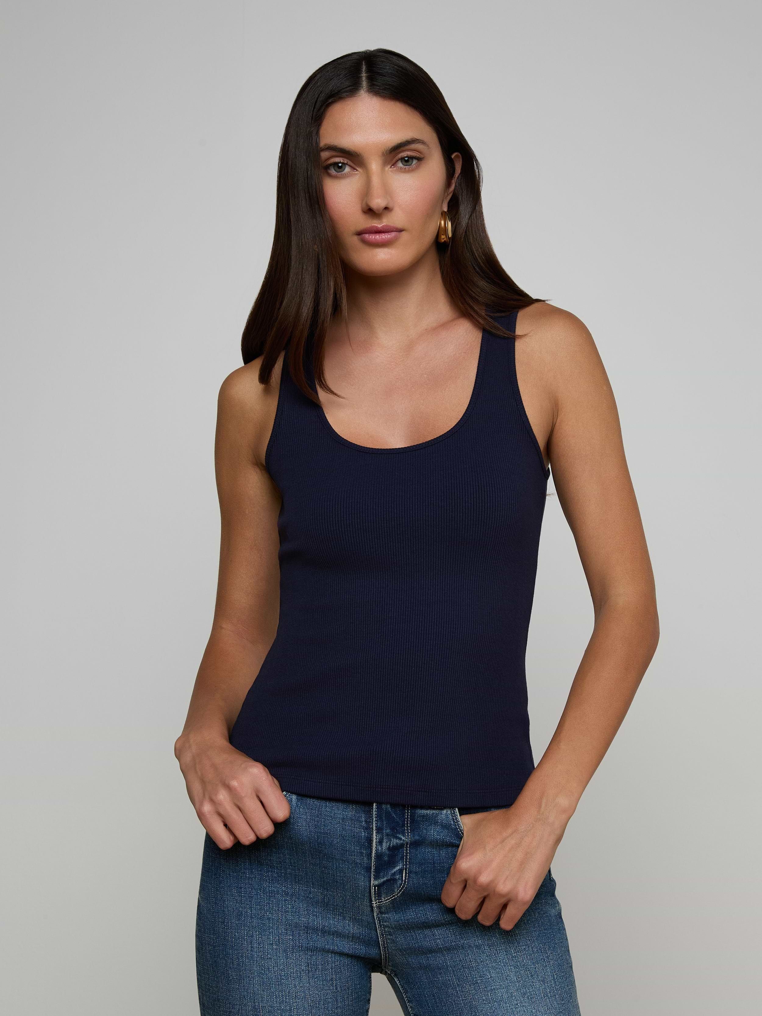 A woman with long dark hair wears the Adira Scoopneck Tank in navy blue and blue jeans, standing against a plain light background, looking at the camera with a neutral expression.