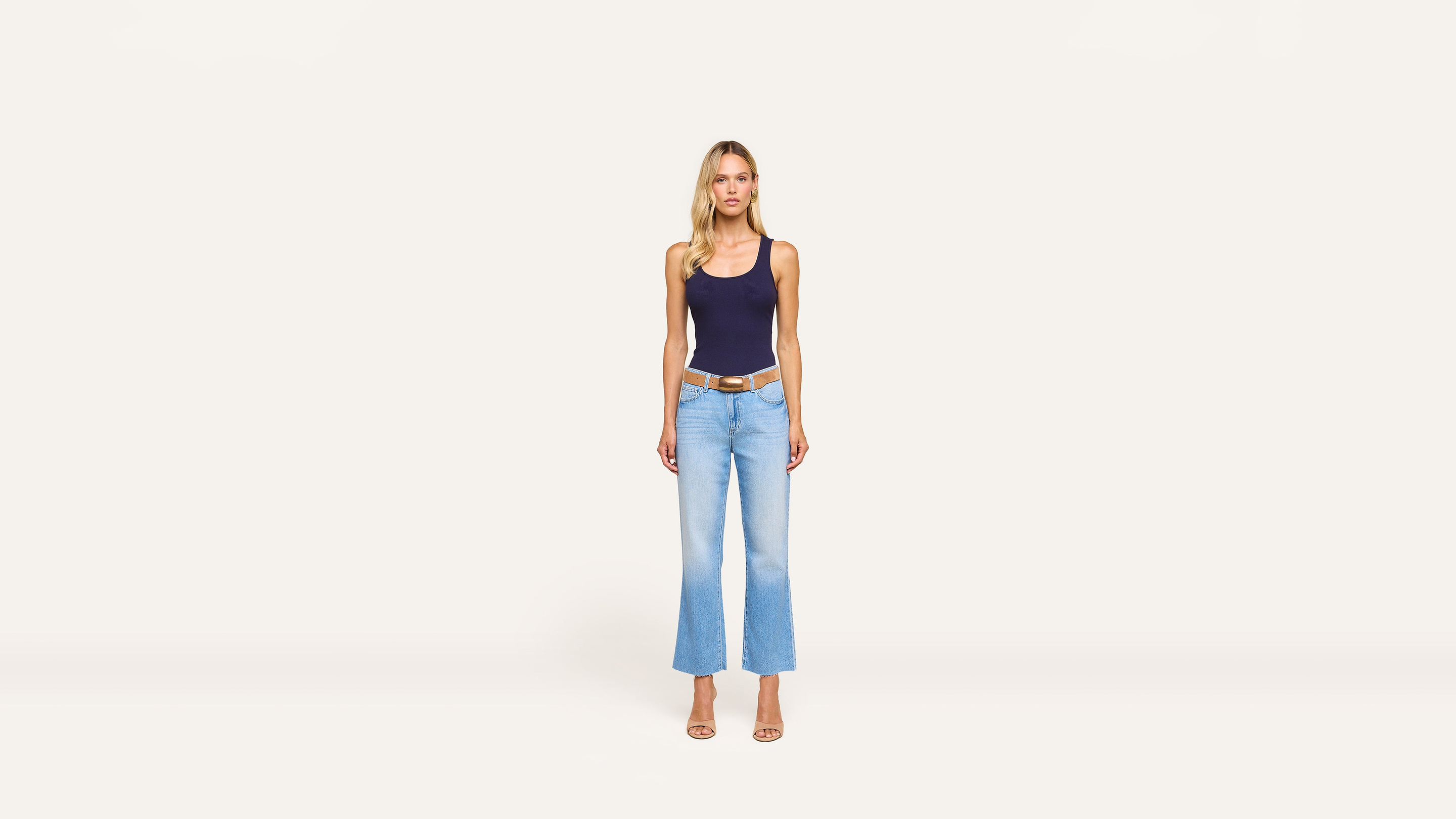 A woman stands against a plain white background, wearing a navy tank top, light blue flared jeans, and beige heels for a chic, casual look.