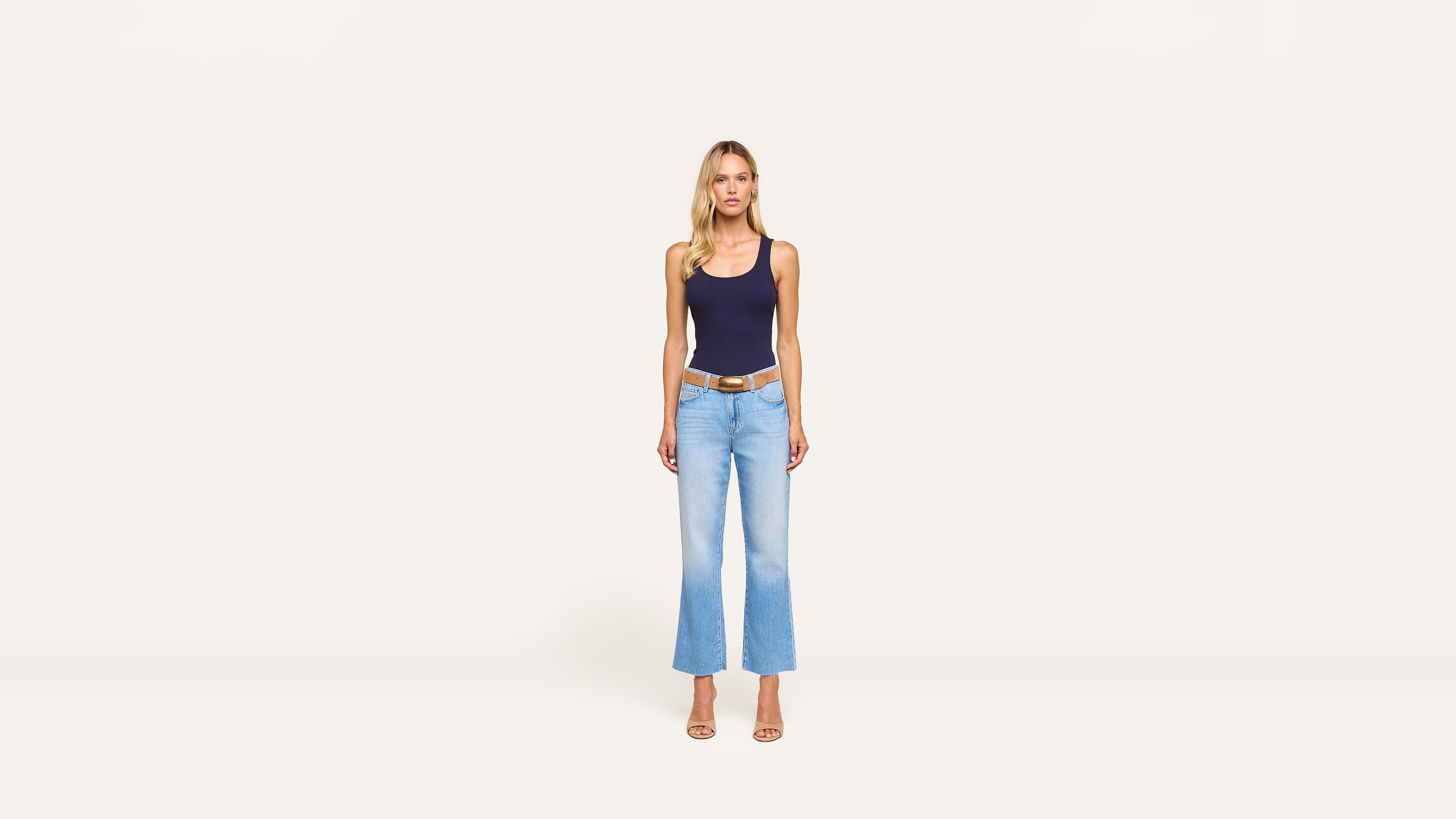A woman stands against a plain white background, wearing a navy tank top, light blue flared jeans, and beige heels for a chic, casual look.