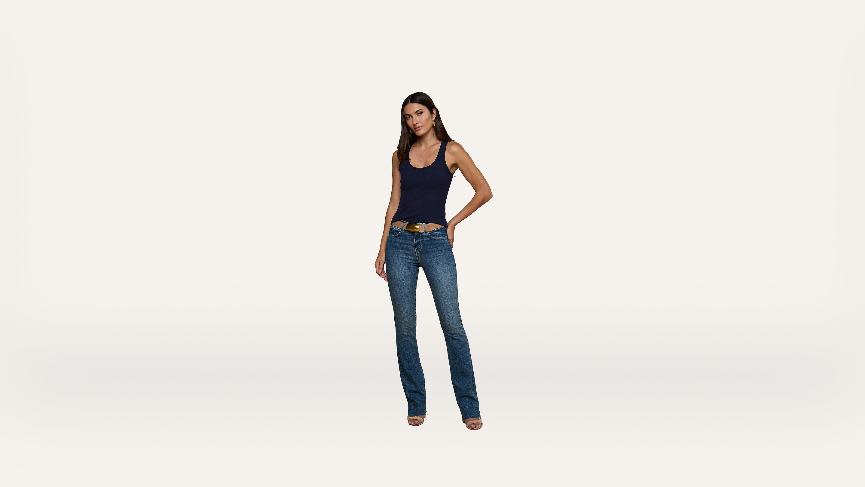 A woman stands facing forward, wearing a navy tank top, blue jeans, and heeled shoes against a plain white background.