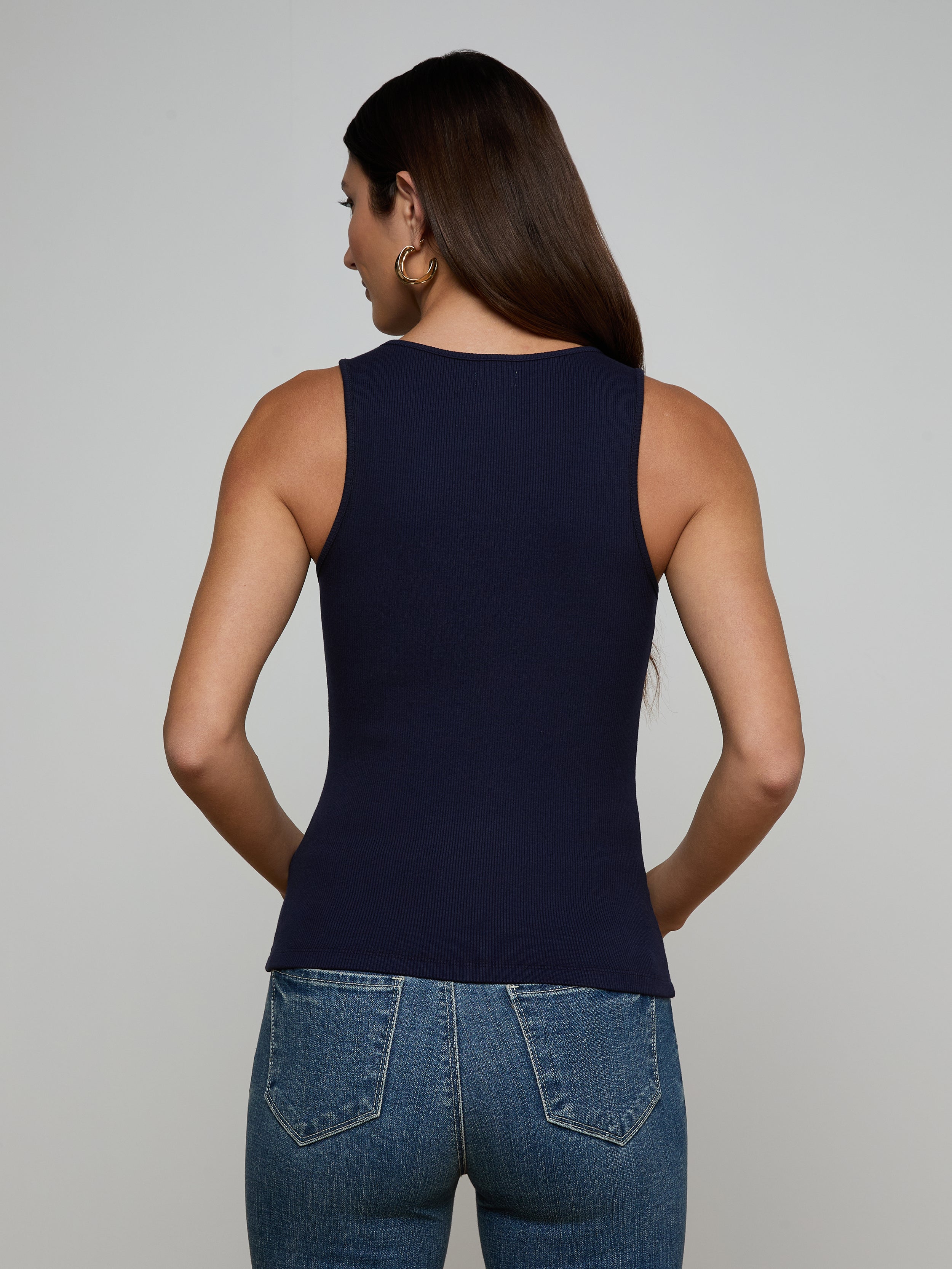 A woman with long dark hair, wearing the Adira Scoopneck Tank and blue jeans, stands facing away from the camera against a plain light background.