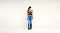 Woman stands against a plain white background, wearing sunglasses, a fitted patterned tank top, wide-leg blue jeans, and brown heeled shoes. She faces forward with a neutral expression.