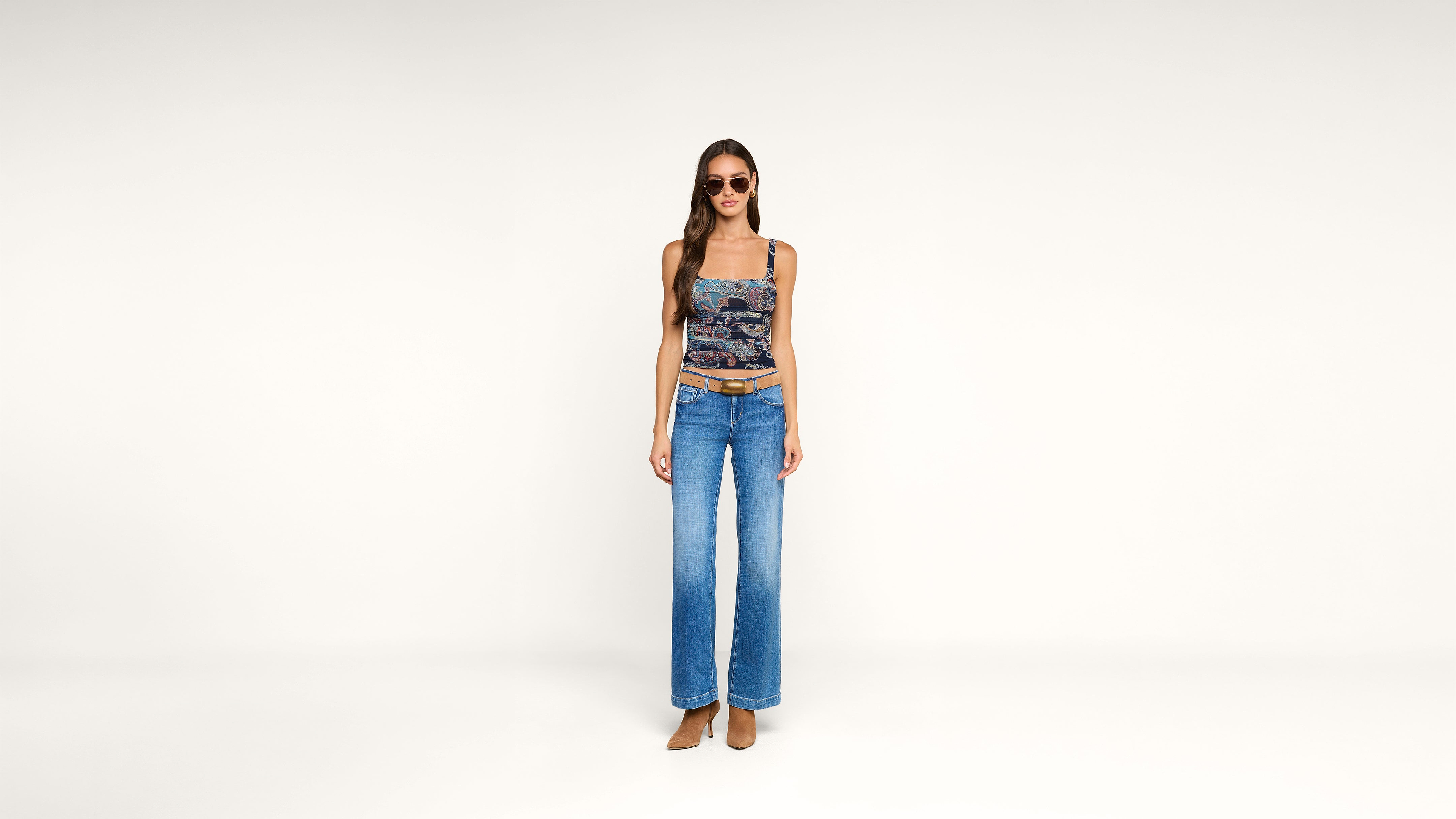 Woman stands against a plain white background, wearing sunglasses, a fitted patterned tank top, wide-leg blue jeans, and brown heeled shoes. She faces forward with a neutral expression.