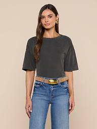 A woman with long brown hair is wearing a dark gray Daria Oversized Cropped Tee, blue jeans with a tan belt, and gold earrings, standing against a plain light background.