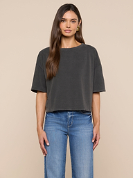 A woman faces forward against a plain off-white background, wearing the Daria Oversized Cropped Tee in black styled with light blue jeans. Her straight brown hair is worn simply, and gold hoop earrings complete her look.