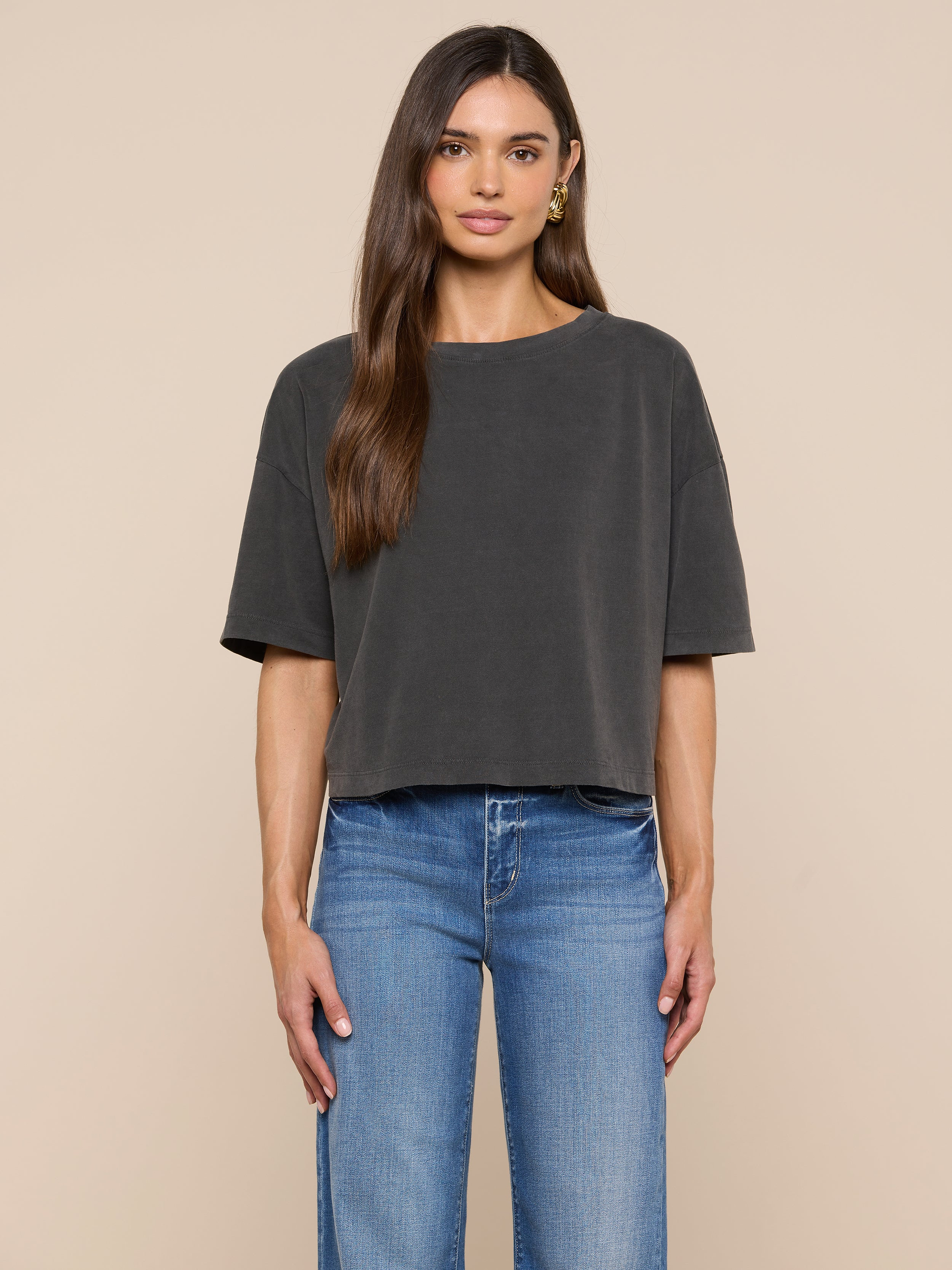 A woman faces forward against a plain off-white background, wearing the Daria Oversized Cropped Tee in black styled with light blue jeans. Her straight brown hair is worn simply, and gold hoop earrings complete her look.