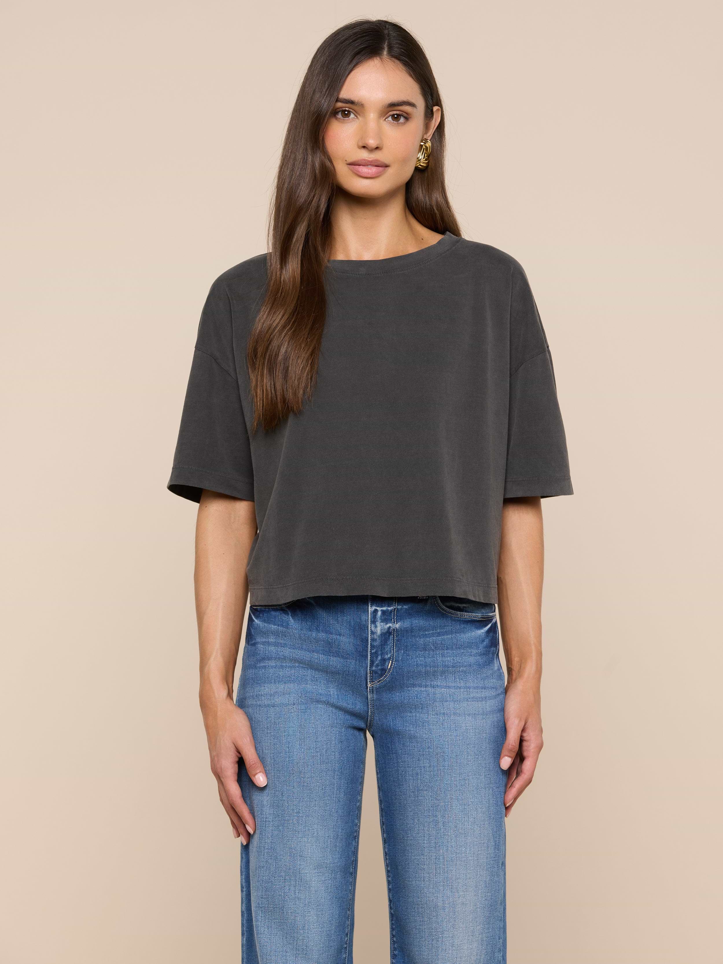 A woman faces forward against a plain off-white background, wearing the Daria Oversized Cropped Tee in black styled with light blue jeans. Her straight brown hair is worn simply, and gold hoop earrings complete her look.