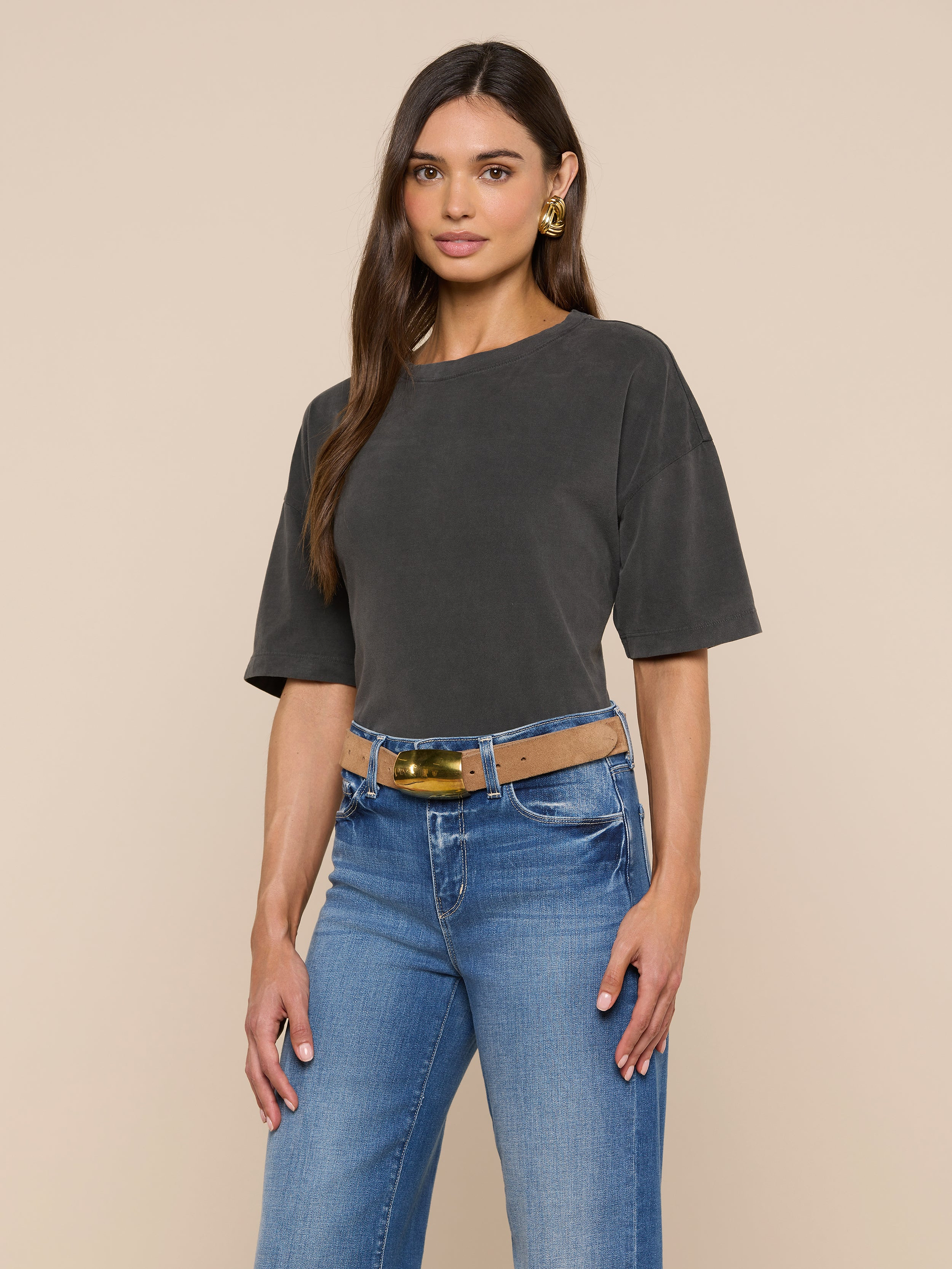 A woman with long brown hair and gold earrings stands against a plain light background, wearing the Daria Oversized Cropped Tee in dark gray. The short-sleeve t-shirt is styled in an oversized fit, tucked into blue jeans with a wide belt.