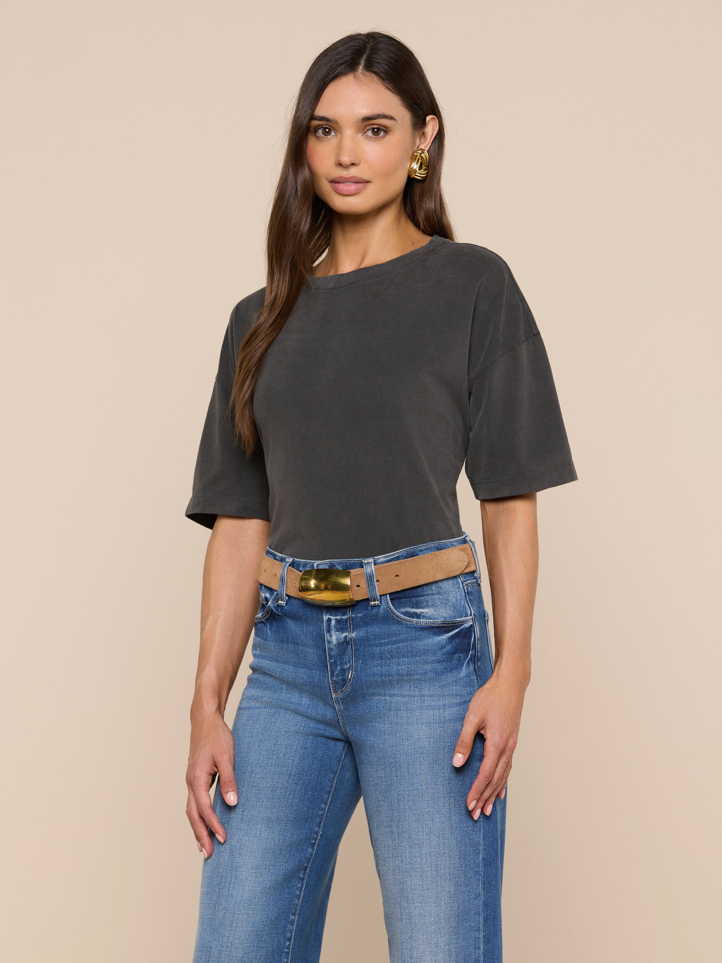 A woman with long brown hair and gold earrings stands against a plain light background, wearing the Daria Oversized Cropped Tee in dark gray. The short-sleeve t-shirt is styled in an oversized fit, tucked into blue jeans with a wide belt.