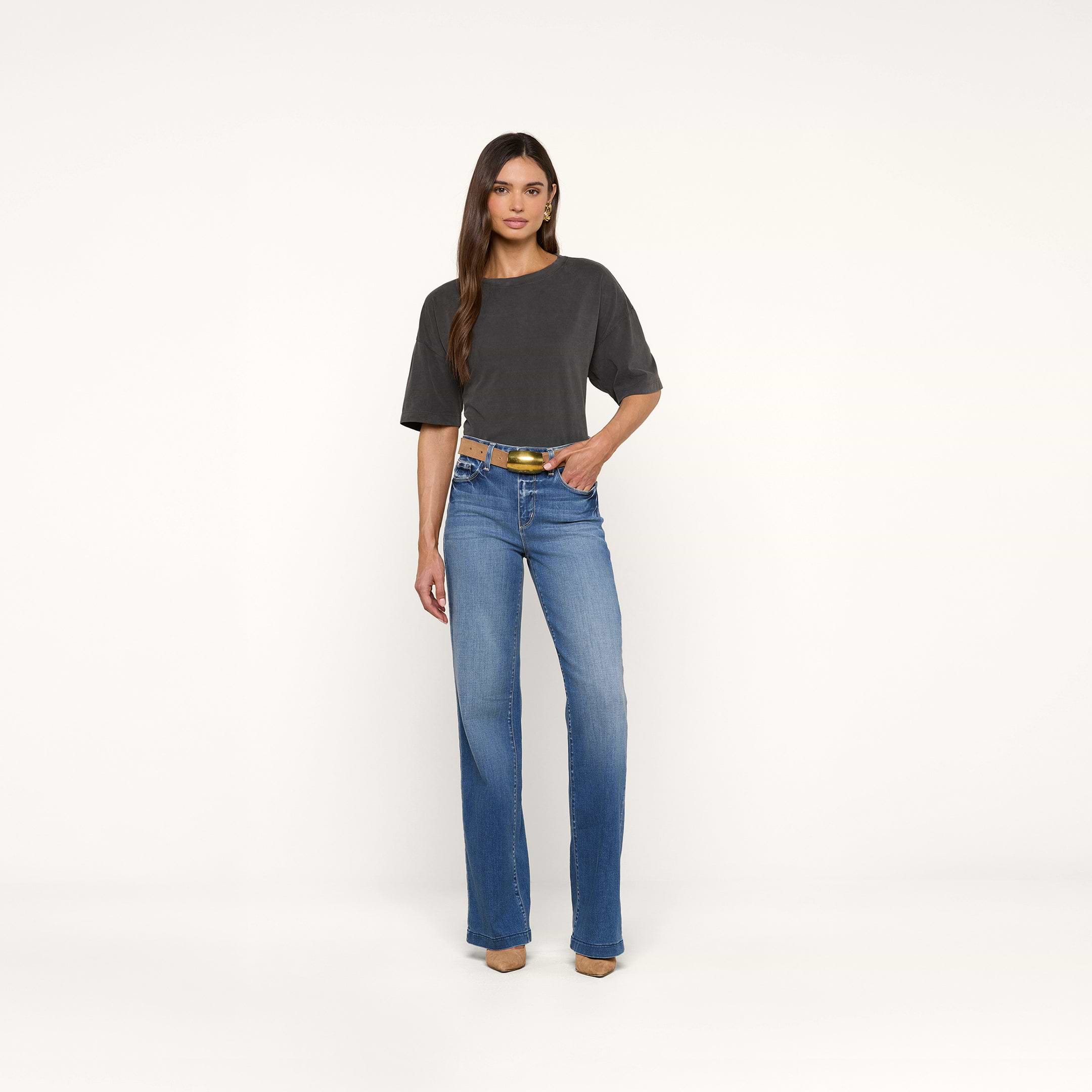 A woman stands against a plain white background wearing the Daria Oversized Cropped Tee in dark gray with blue jeans, a yellow belt, and tan shoes. She has long, straight hair and one hand in her pocket.
