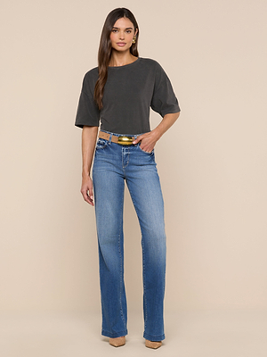 A woman with long hair and a neutral expression stands against a beige background wearing an oversized dark gray Daria Oversized Cropped Tee, light blue high-waisted jeans, a tan belt, and tan pointed shoes.