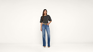 Woman stands against a plain white background, wearing a dark gray t-shirt, blue jeans, and beige shoes. She has long, straight brown hair and one hand in her pocket.
