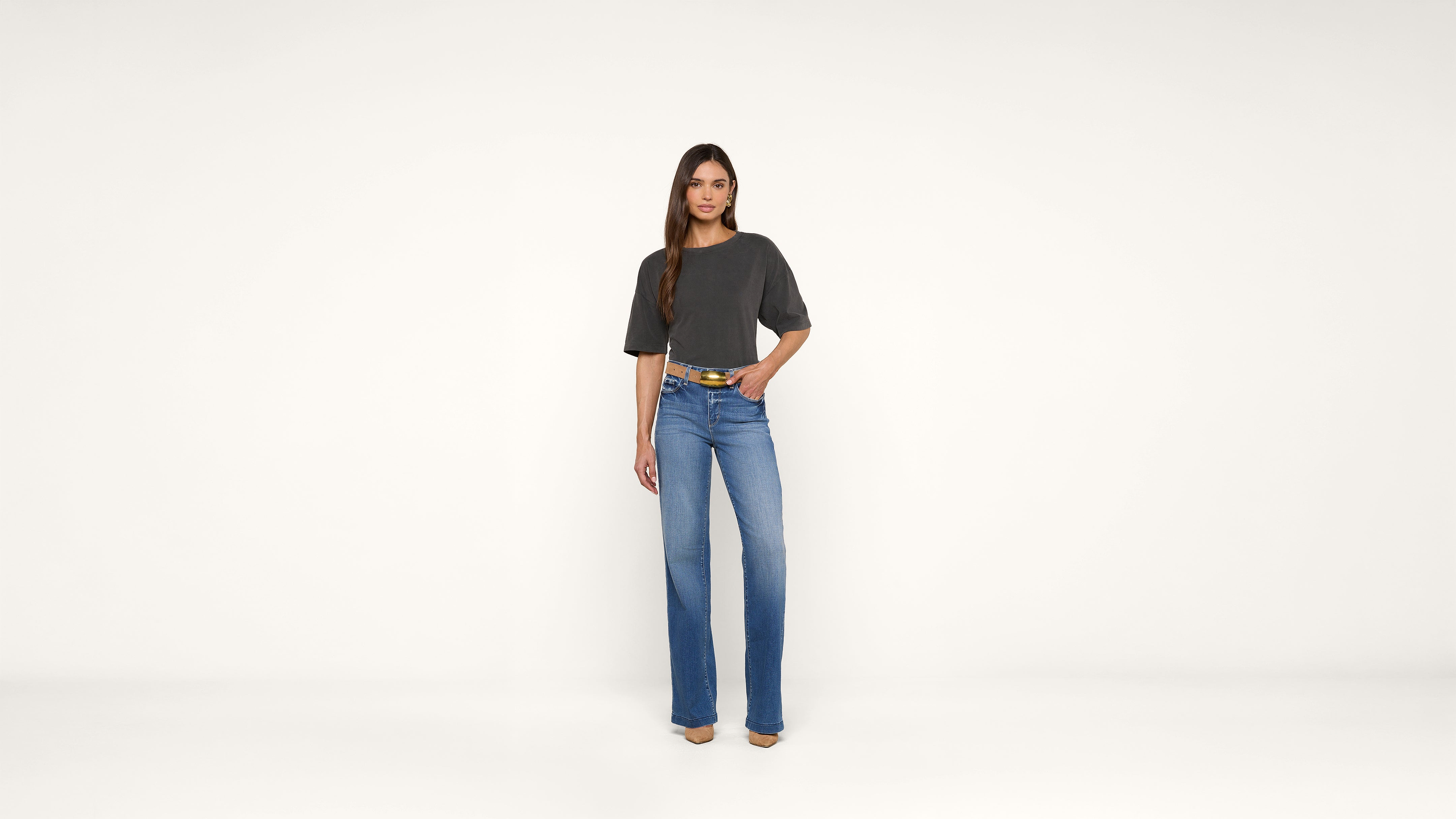Woman stands against a plain white background, wearing a dark gray t-shirt, blue jeans, and beige shoes. She has long, straight brown hair and one hand in her pocket.