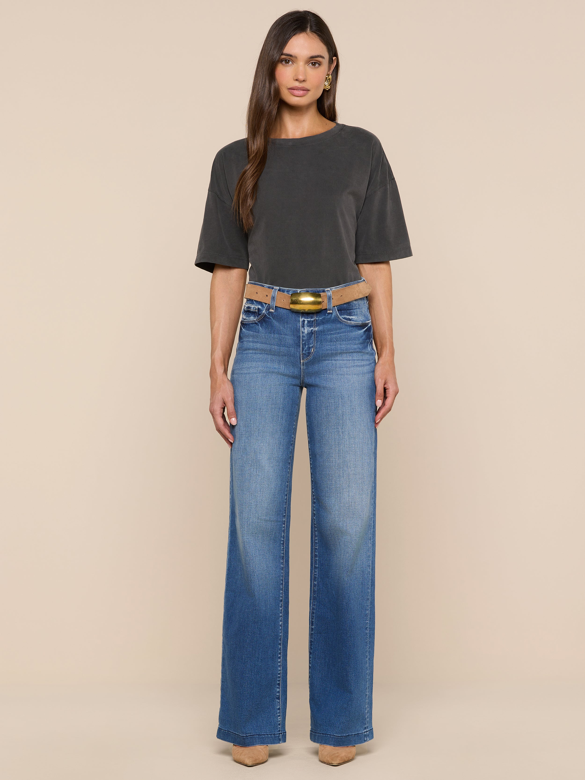 A woman stands facing forward, wearing the Daria Oversized Cropped Tee in dark gray with an oversized fit, blue wide-leg jeans, a black belt with a gold buckle, and beige pointed shoes against a plain white background.
