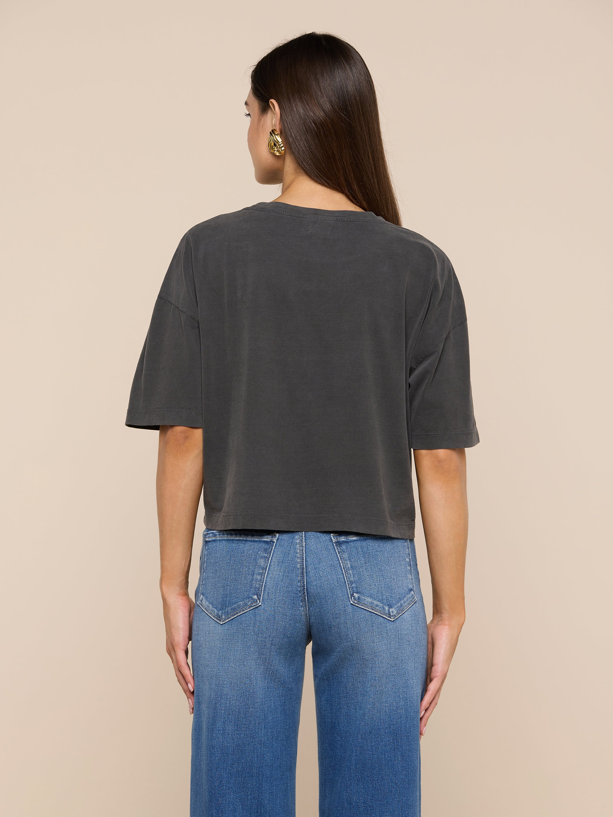 A woman stands facing away, wearing the Daria Oversized Cropped Tee in dark gray, made from Supima cotton, paired with blue jeans. Her straight brown hair falls over her shoulders, and she accessorizes with gold hoop earrings. The background is plain and light-colored.