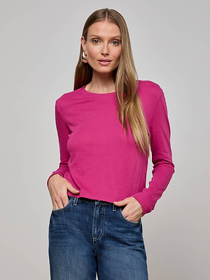 A woman with long straight hair wears the bright pink Zeva Tee, made from organic cotton, and blue jeans. She stands facing the camera with her hands in her pockets against a plain light gray background.
