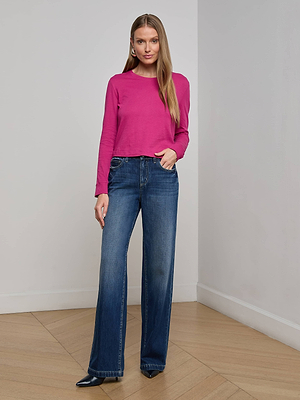 A woman stands indoors wearing the Zeva Tee, a bright pink organic cotton long-sleeve top, paired with blue wide-leg jeans and black pointed-toe shoes. She poses with one hand in her pocket against a plain background.