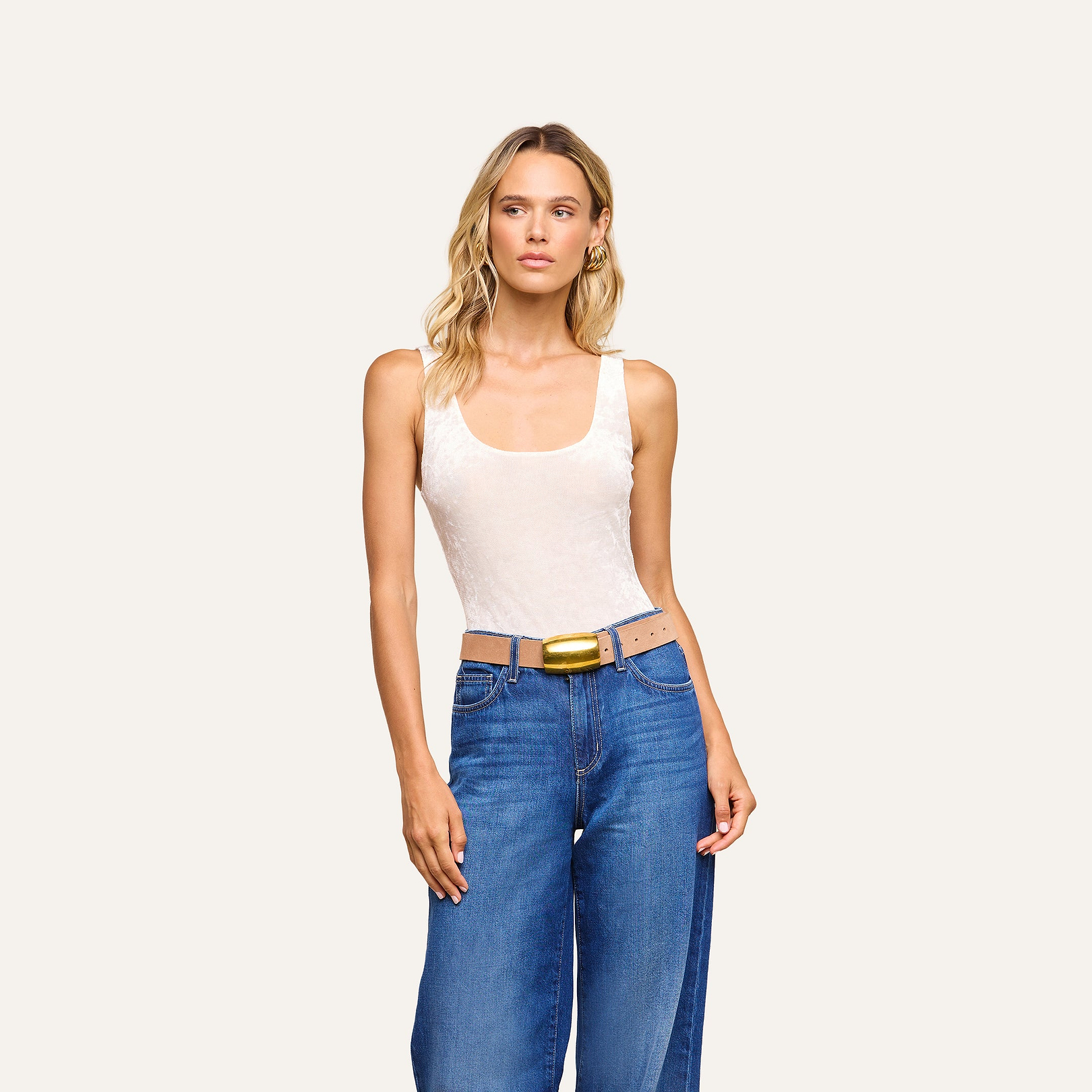 A woman with wavy blonde hair wears the Kalea Velvet Bodysuit in ivory, paired with wide blue jeans and a tan belt featuring a large yellow buckle, standing against a plain light background.