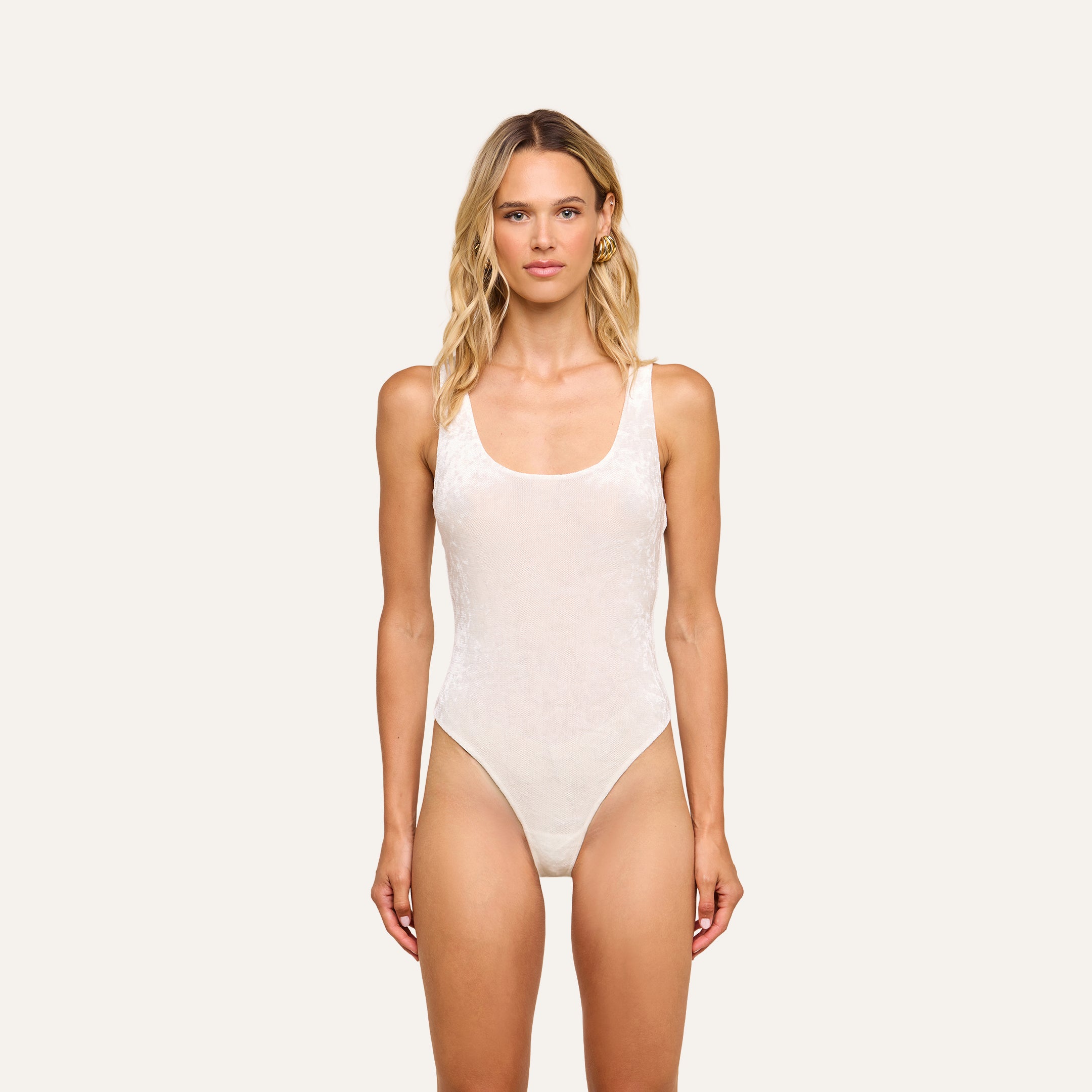 A woman with wavy blonde hair faces forward, wearing the Kalea Velvet Bodysuit in ivory as a lightweight layer against a plain, light background.
