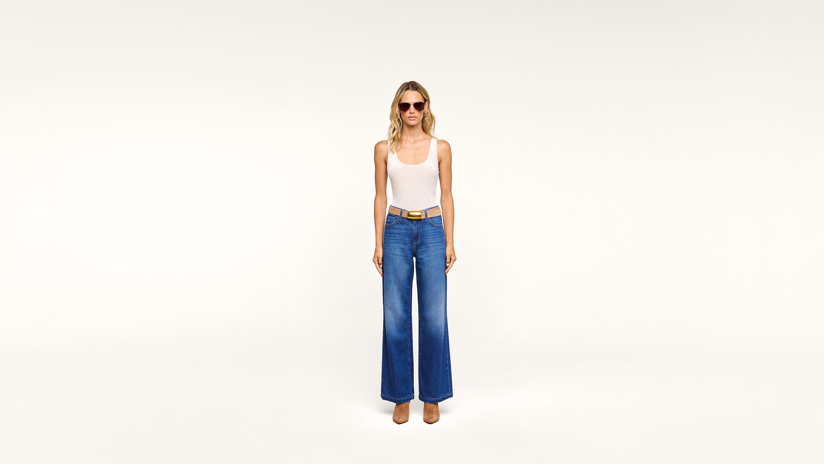 Woman wearing sunglasses, a white tank top, wide-leg blue jeans, and tan shoes stands against a plain white background, facing forward with arms relaxed at her sides.