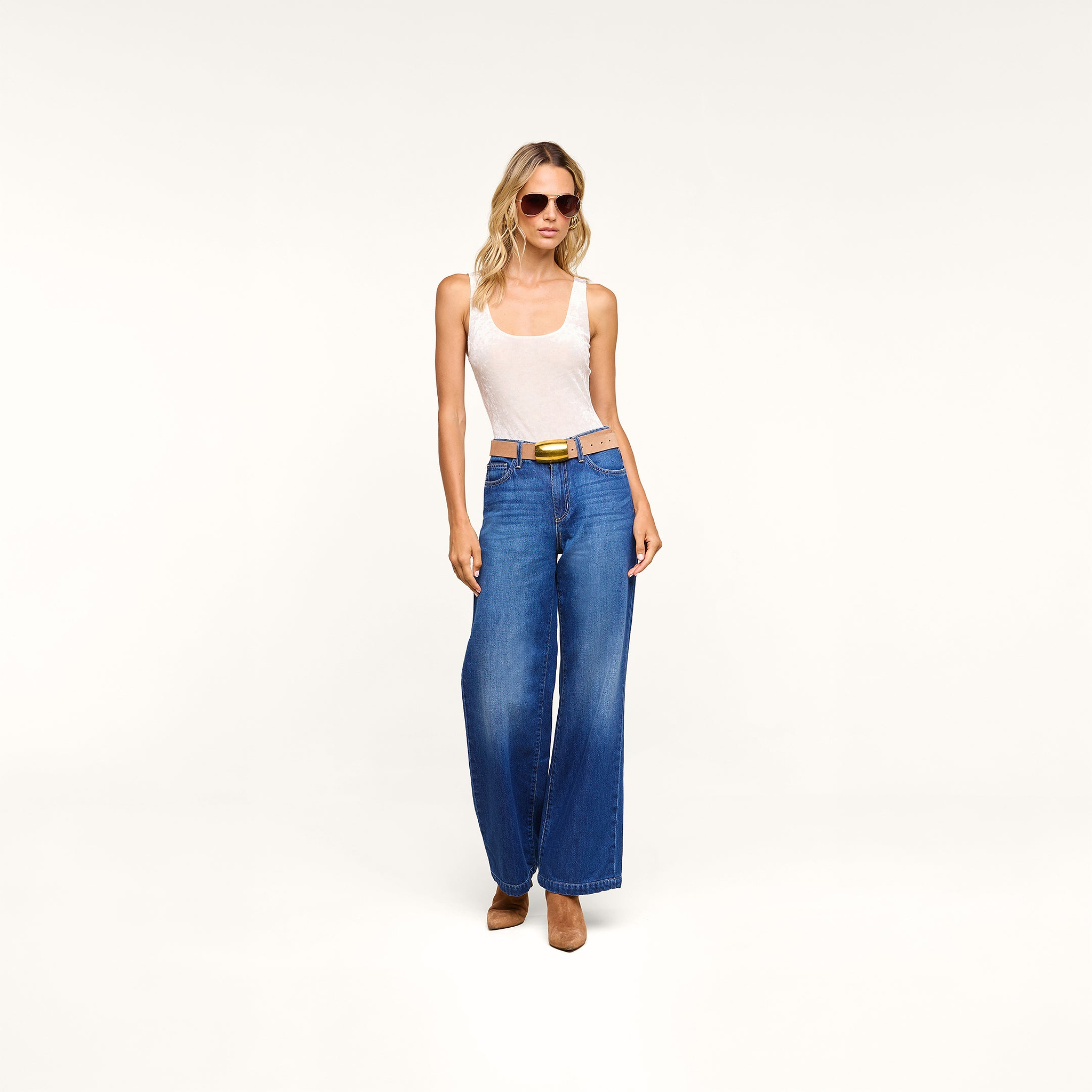 Woman wearing sunglasses, a Kalea Velvet Bodysuit styled as a white tank top, blue wide-leg jeans with a belt, and brown heels stands confidently against a plain white background.