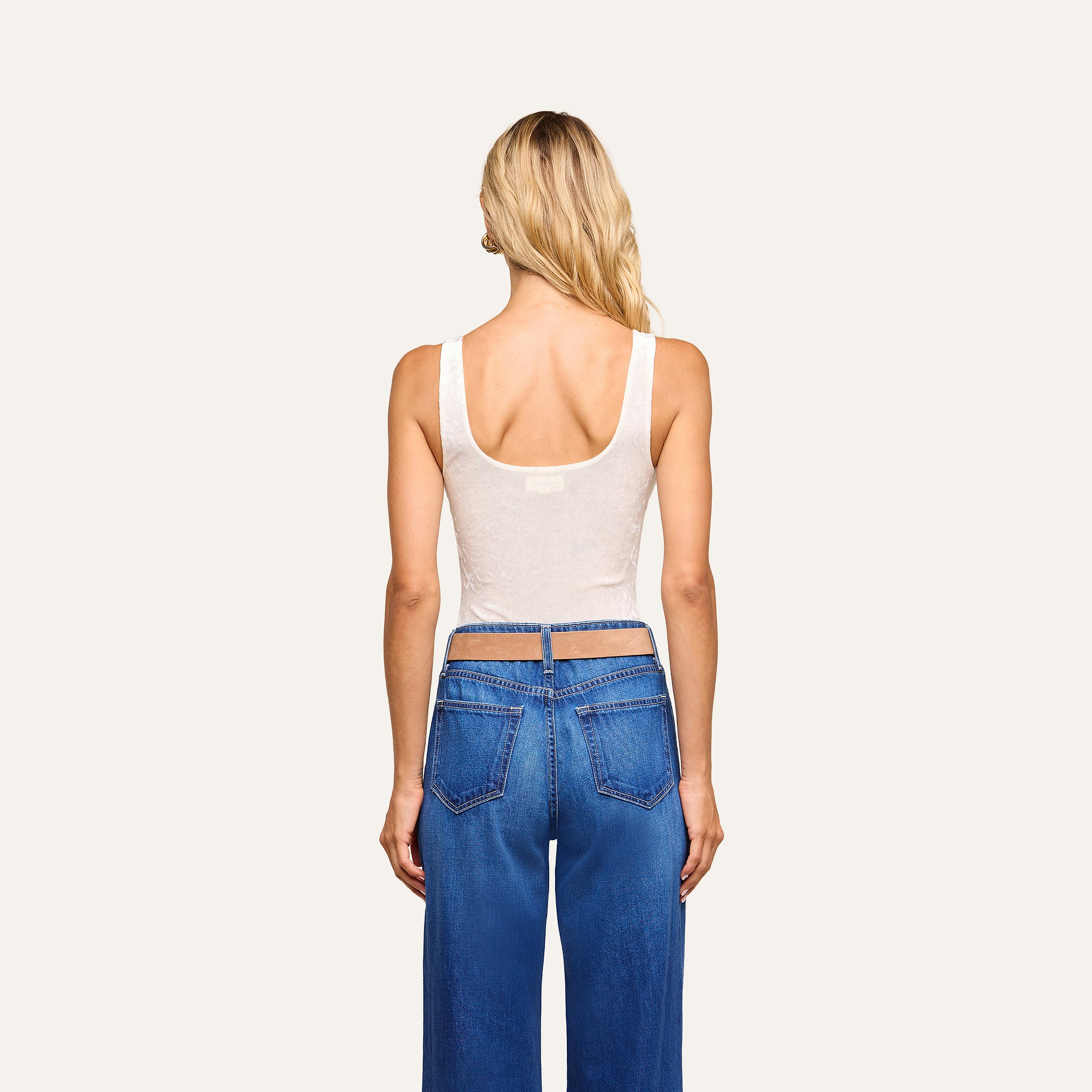 A woman with long blonde hair stands facing away from the camera against a plain light background, wearing blue jeans and the Kalea Velvet Bodysuit styled as a lightweight layer.