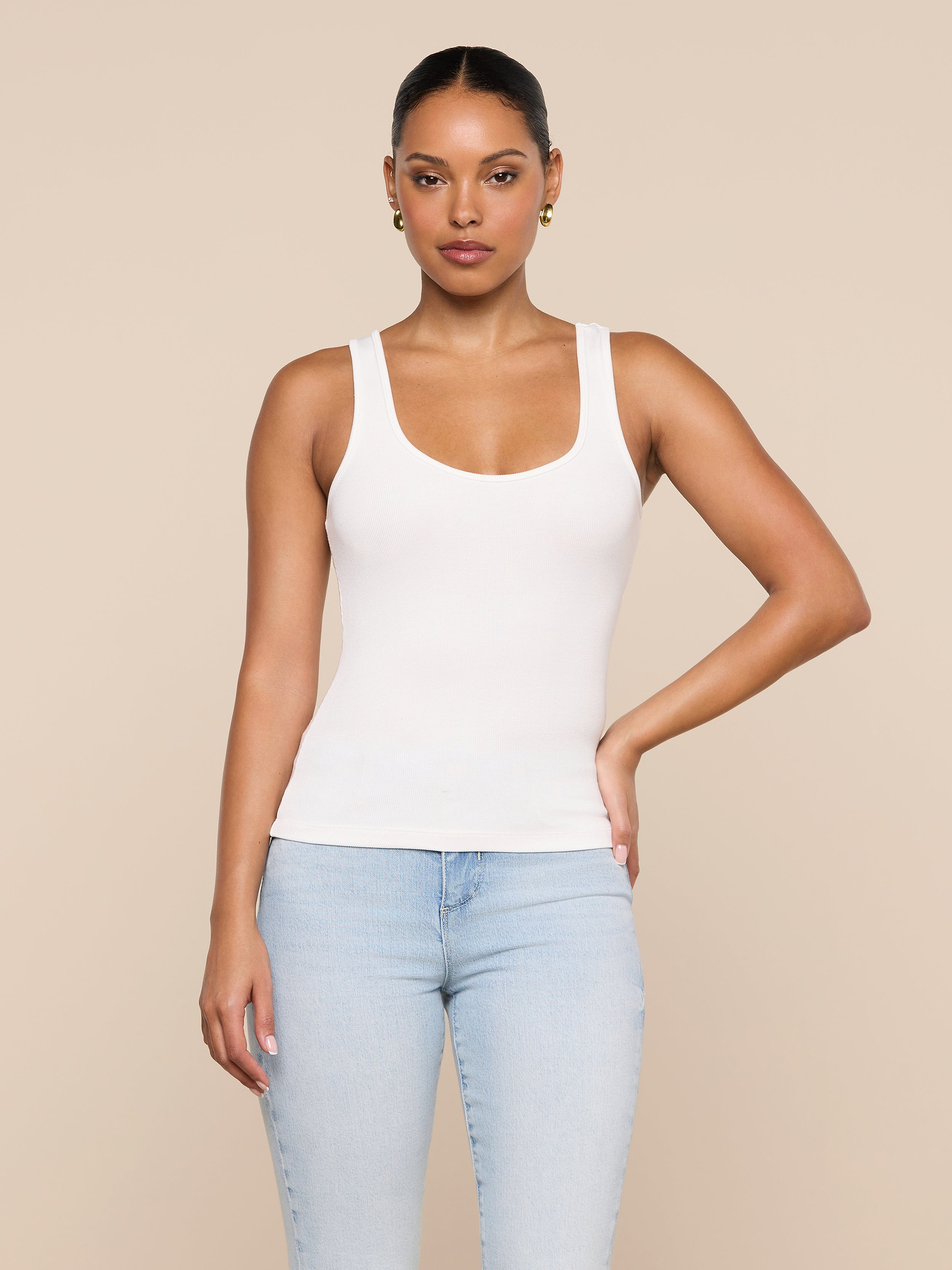 A woman stands against a plain background wearing the Cole Scoopneck Tank and light blue jeans, one arm resting on her hip and maintaining a neutral facial expression.