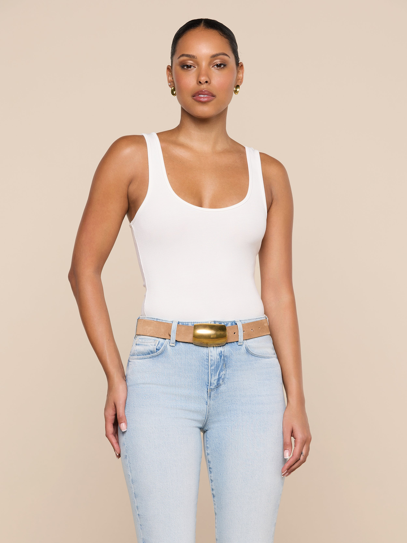 A woman wearing the Cole Scoopneck Tank in white, paired with light blue jeans featuring cutouts at the waist and a gold buckle belt, stands against a plain light background.