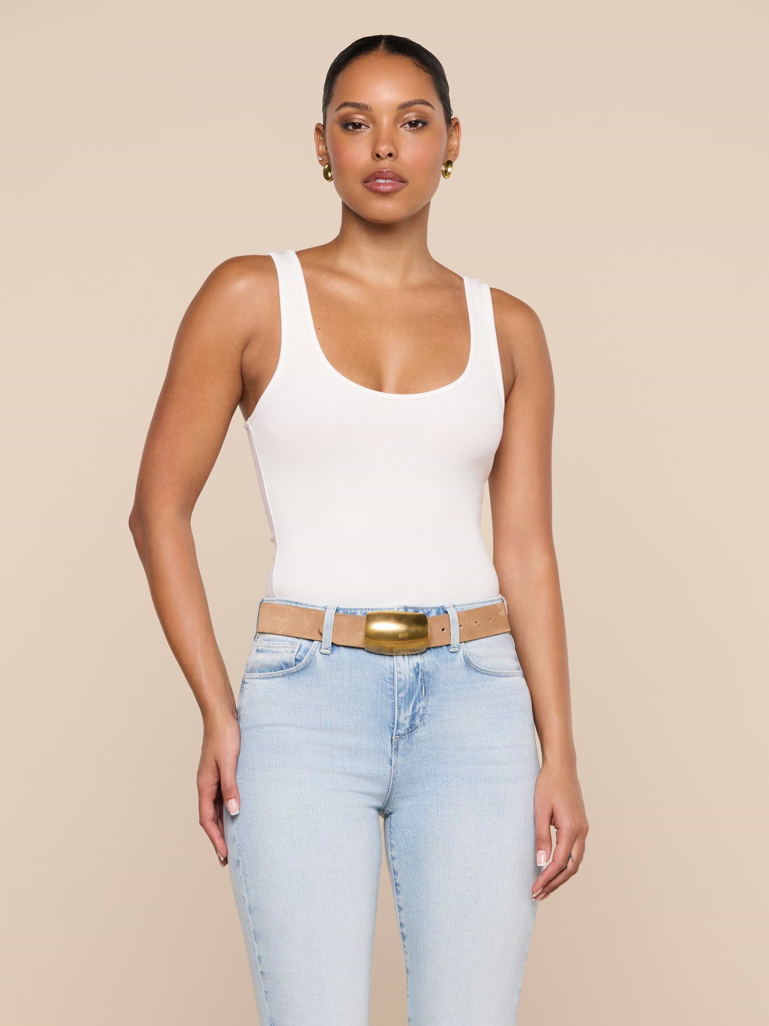 A woman wearing the Cole Scoopneck Tank in white, paired with light blue jeans featuring cutouts at the waist and a gold buckle belt, stands against a plain light background.
