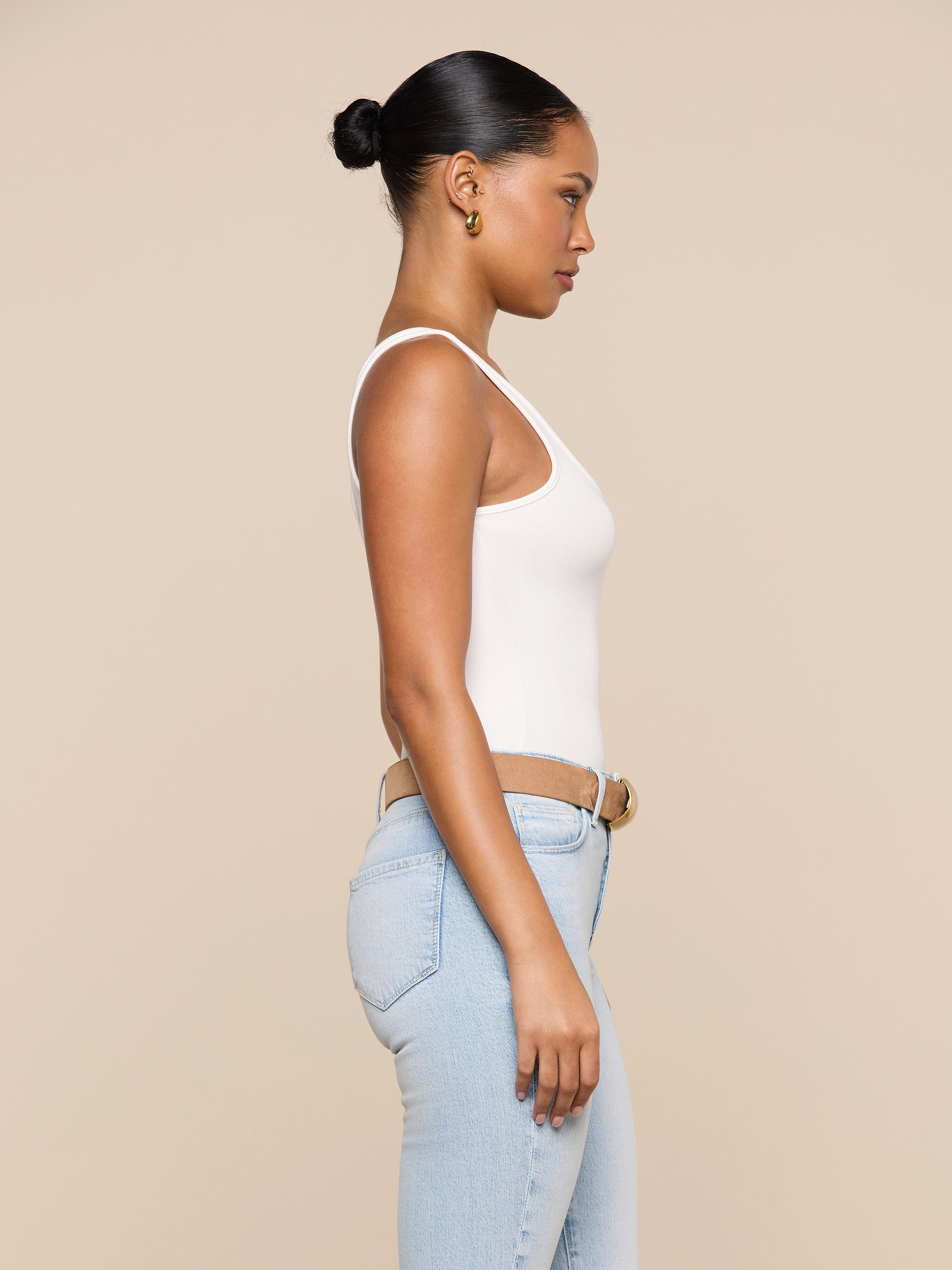 A woman wears the Cole Scoopneck Tank in white paired with light blue jeans, standing in profile against a plain, light background. Her hair is pulled back in a bun and she gazes straight ahead with a neutral expression.