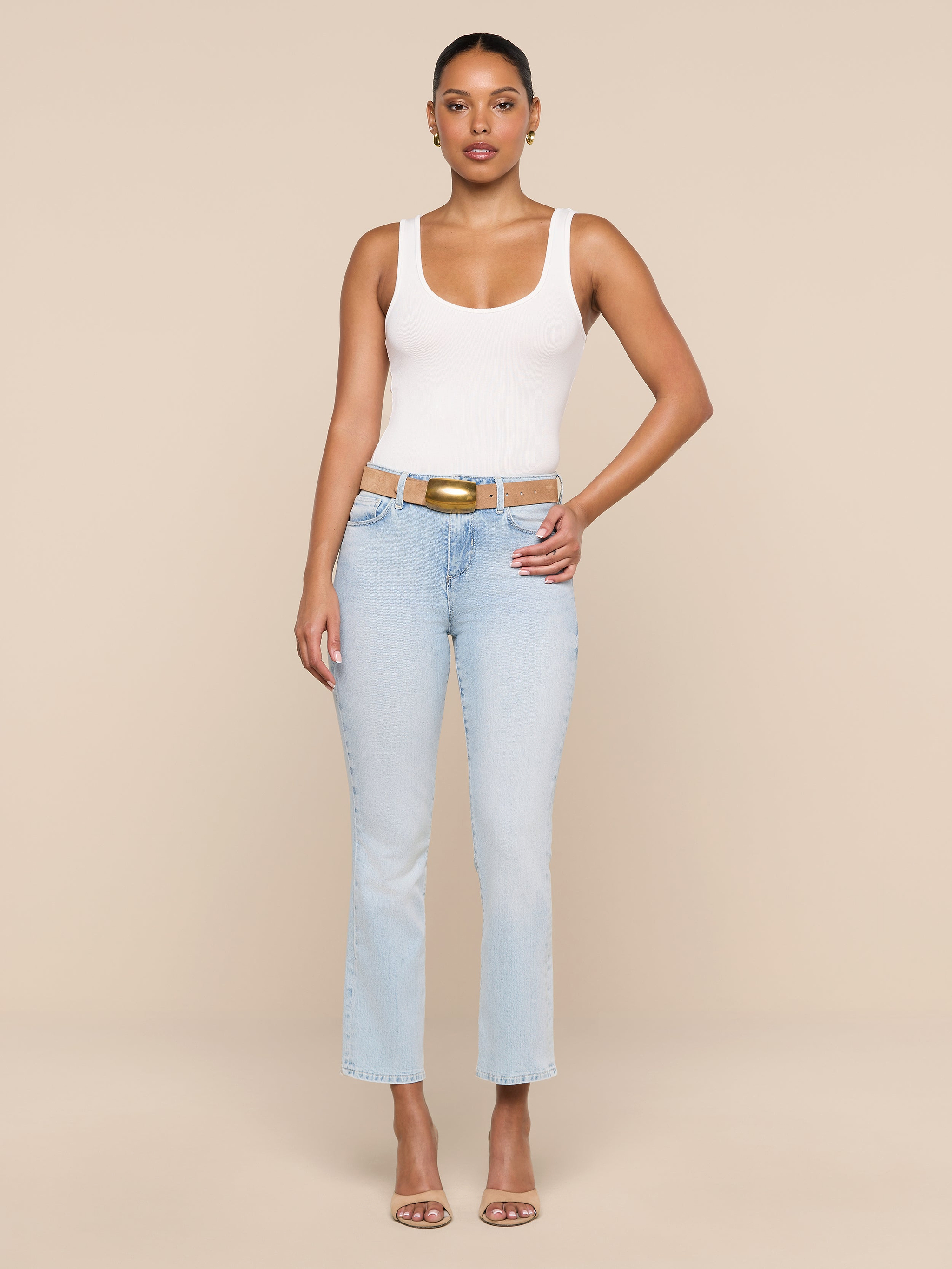A woman stands facing forward against a plain white background, wearing the Cole Scoopneck Tank, light blue jeans, a tan belt, and nude heels. One hand rests on her hip while the other hangs by her side.