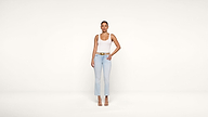 A person stands facing forward against a plain white background, wearing a white tank top, light blue jeans, a brown belt, and heeled sandals, with one hand on their hip.