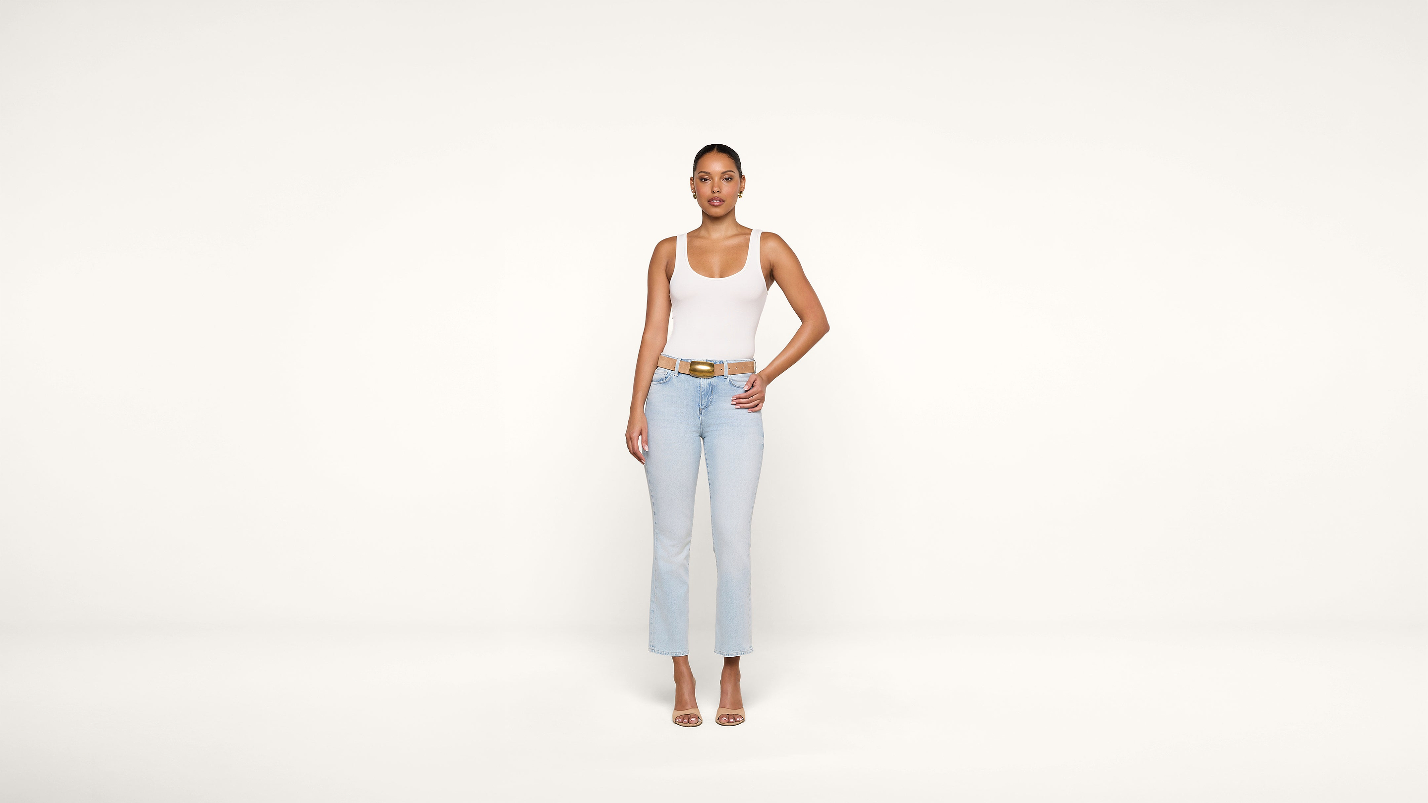 A person stands facing forward against a plain white background, wearing a white tank top, light blue jeans, a brown belt, and heeled sandals, with one hand on their hip.