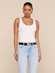 A woman stands against a plain background, wearing the Nyra Bodysuit—a white tank top crafted from soft stretch rib—paired with light blue jeans, a black belt, and hoop earrings. Her hair is pulled back and she has a neutral expression, with her arms relaxed at her sides.