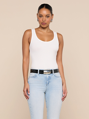 A woman poses against a beige background wearing the white Nyra Bodysuit with light blue jeans and a black belt. Her hair is pulled back, she wears earrings, and gazes directly at the camera with a neutral expression.