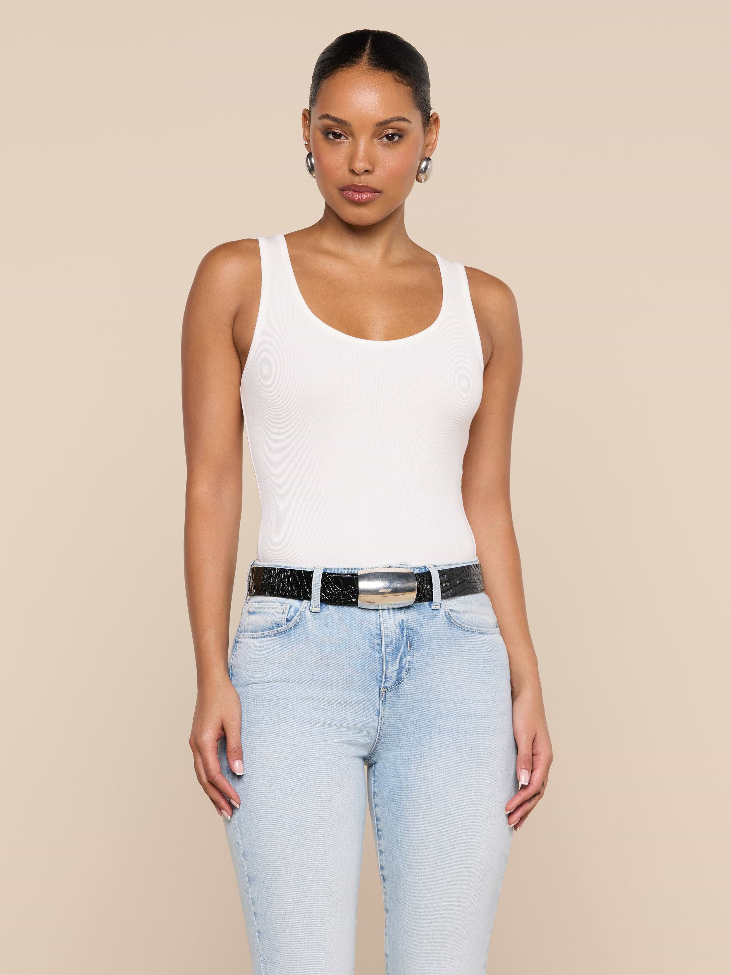 A woman stands against a plain background, wearing the Nyra Bodysuit—a white tank top crafted from soft stretch rib—paired with light blue jeans, a black belt, and hoop earrings. Her hair is pulled back and she has a neutral expression, with her arms relaxed at her sides.