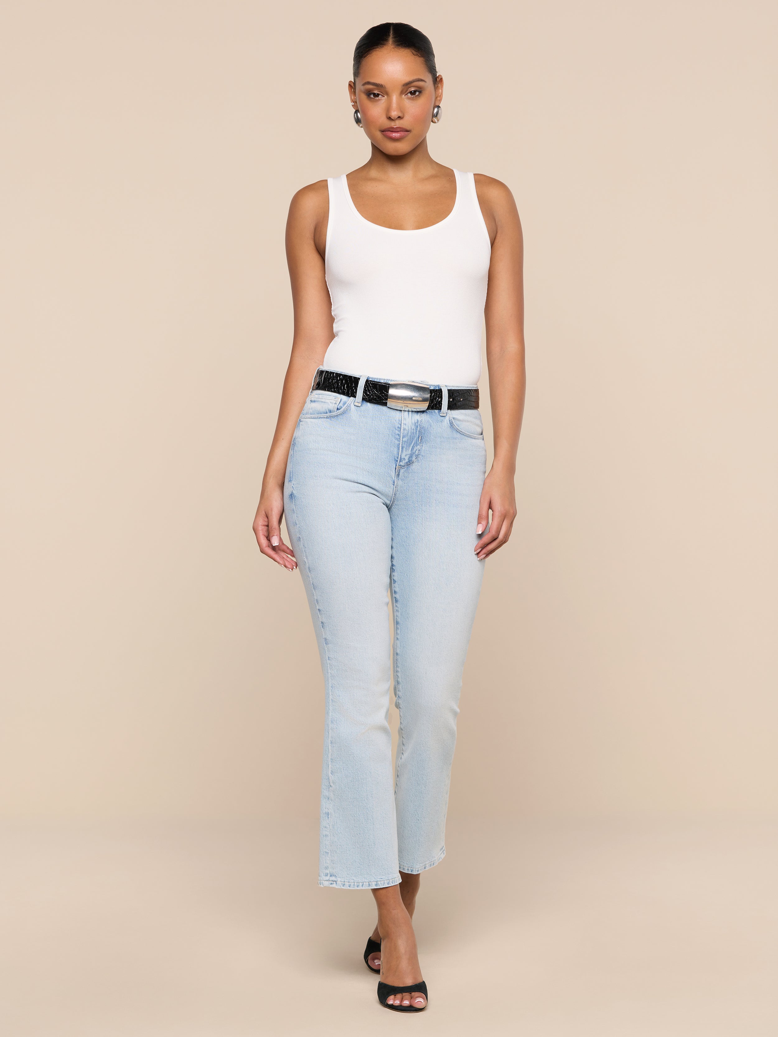 A person wearing the Nyra Bodysuit in white as a base layer, paired with light blue jeans, a black belt, and black heels, stands against a plain white background facing forward with a neutral expression.