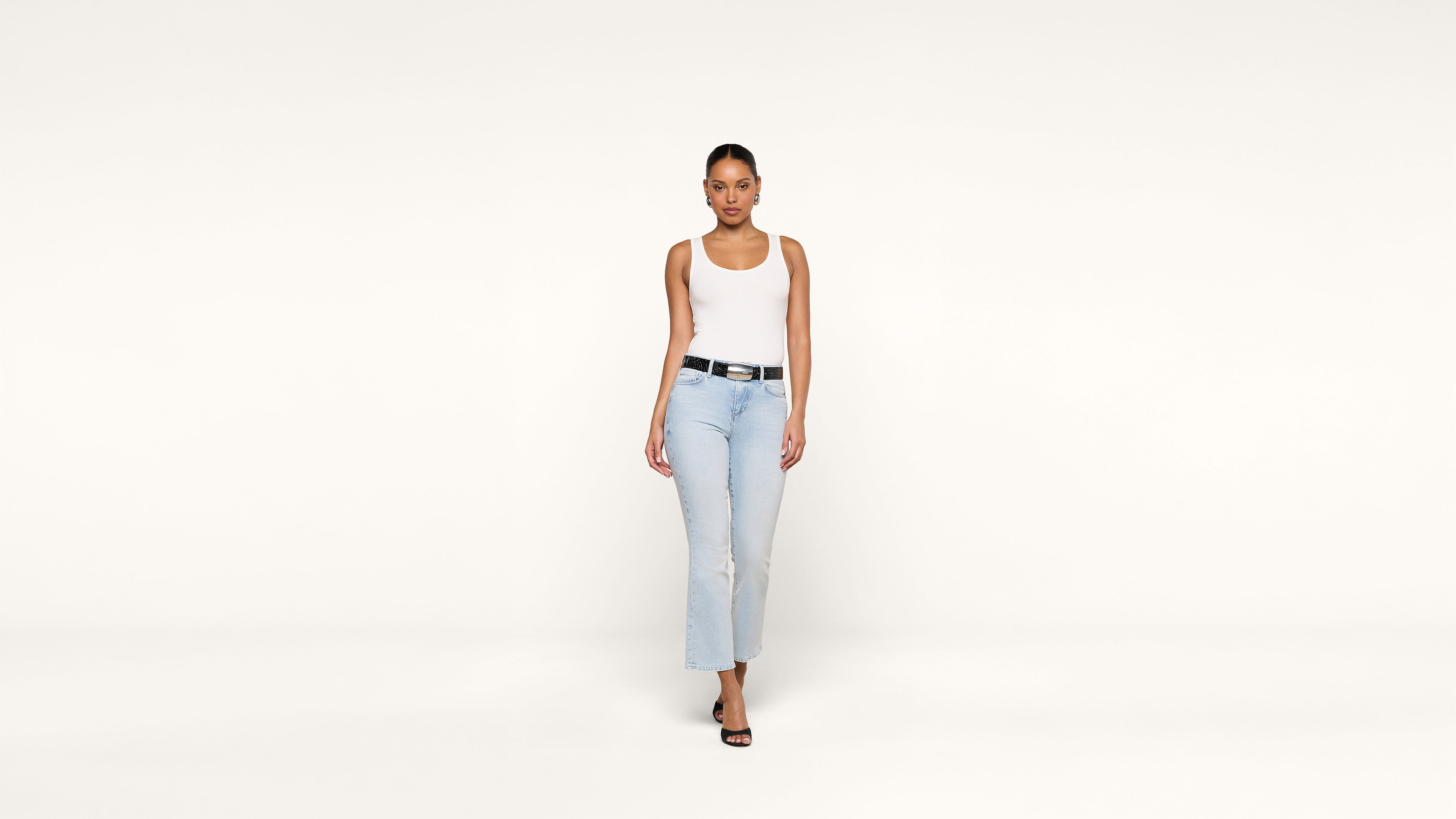 A woman wearing a white tank top, light blue jeans, and black sandals stands against a plain white background, looking directly at the camera.