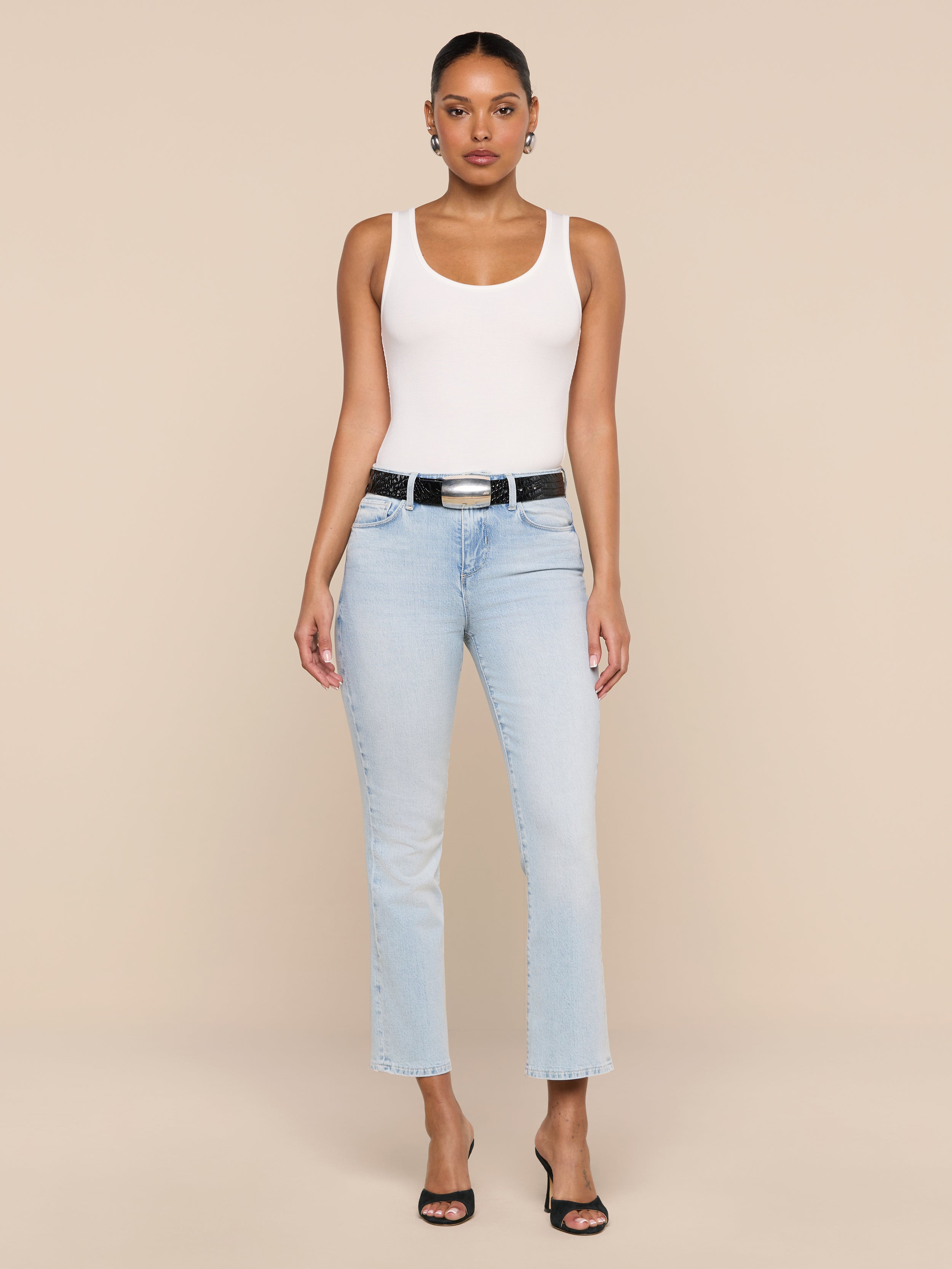 A woman stands facing forward wearing the Nyra Bodysuit as a soft stretch rib base layer, paired with light blue jeans, a black belt, and black open-toe heels against a plain white background.