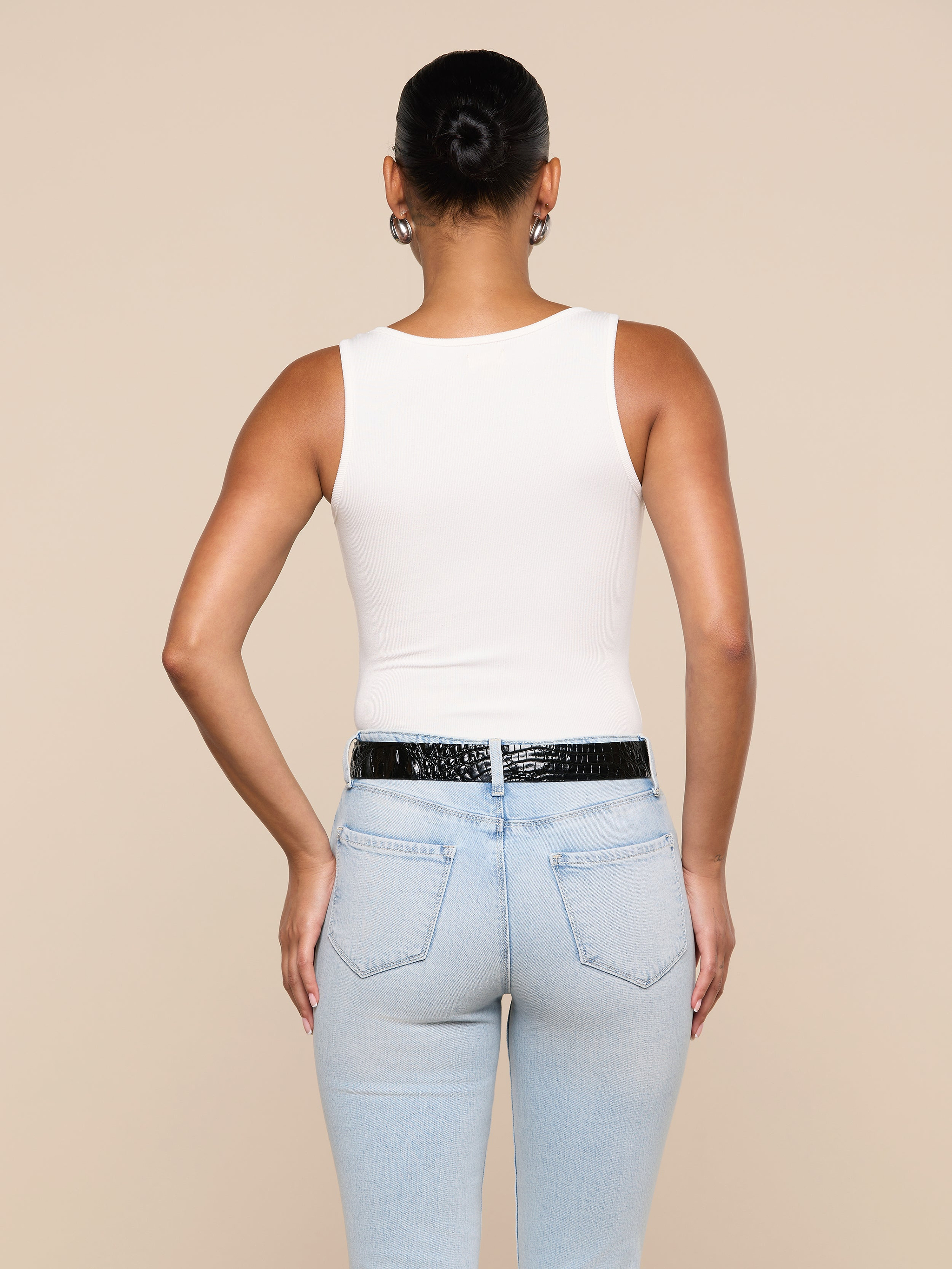 A person with their hair in a bun is seen from the back, wearing the Nyra Bodysuit—a white sleeveless soft stretch rib top—paired with light blue jeans and a black belt, standing against a plain light background.