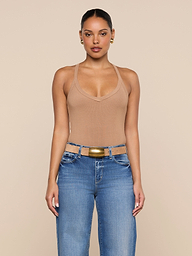 A woman stands facing forward against a plain light background, wearing the Wrenna Racerback Bodysuit in tan rib fabric, blue jeans, a tan belt with a gold buckle, and gold hoop earrings.