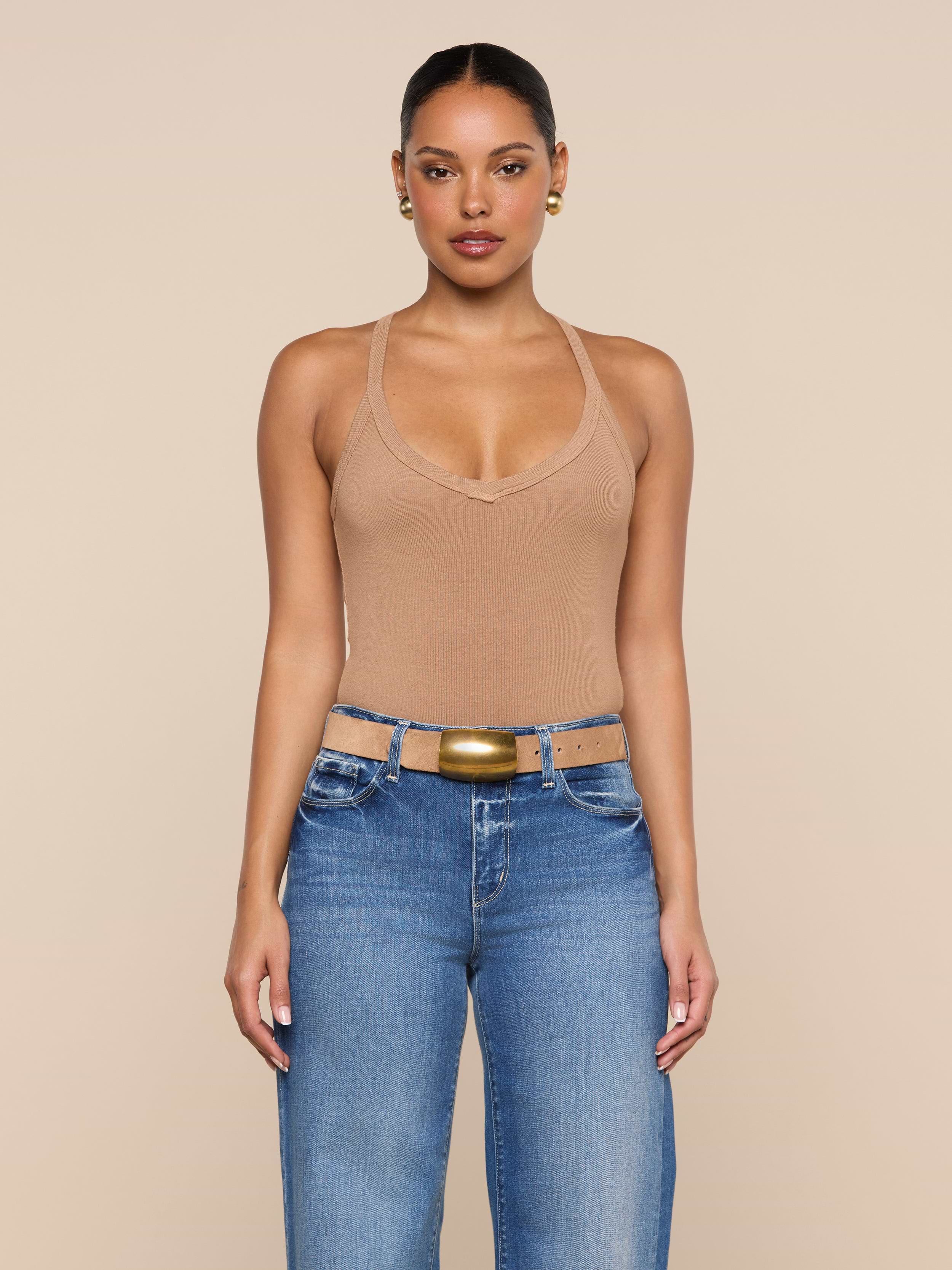 A woman stands facing forward against a plain light background, wearing the Wrenna Racerback Bodysuit in tan rib fabric, blue jeans, a tan belt with a gold buckle, and gold hoop earrings.