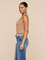 A woman stands in profile against a plain background, wearing the Wrenna Racerback Bodysuit in tan rib fabric, paired with blue jeans and a beige belt; her hair is pulled back and she accessorizes with gold hoop earrings.