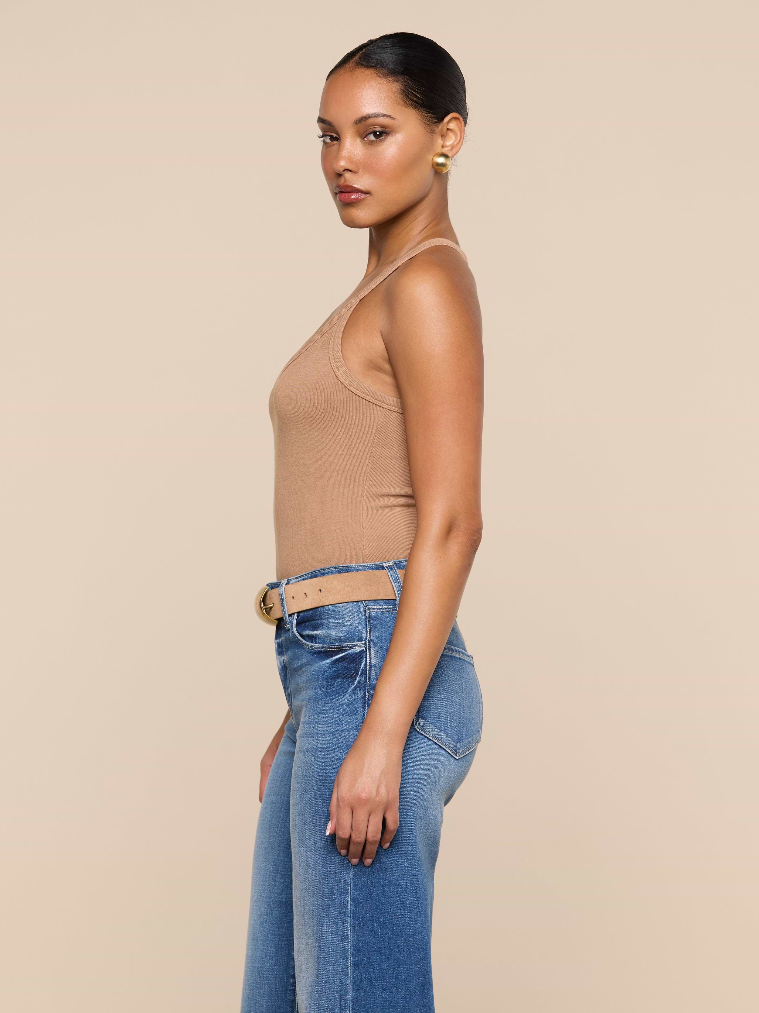 A woman stands in profile against a plain background, wearing the Wrenna Racerback Bodysuit in tan rib fabric, paired with blue jeans and a beige belt; her hair is pulled back and she accessorizes with gold hoop earrings.