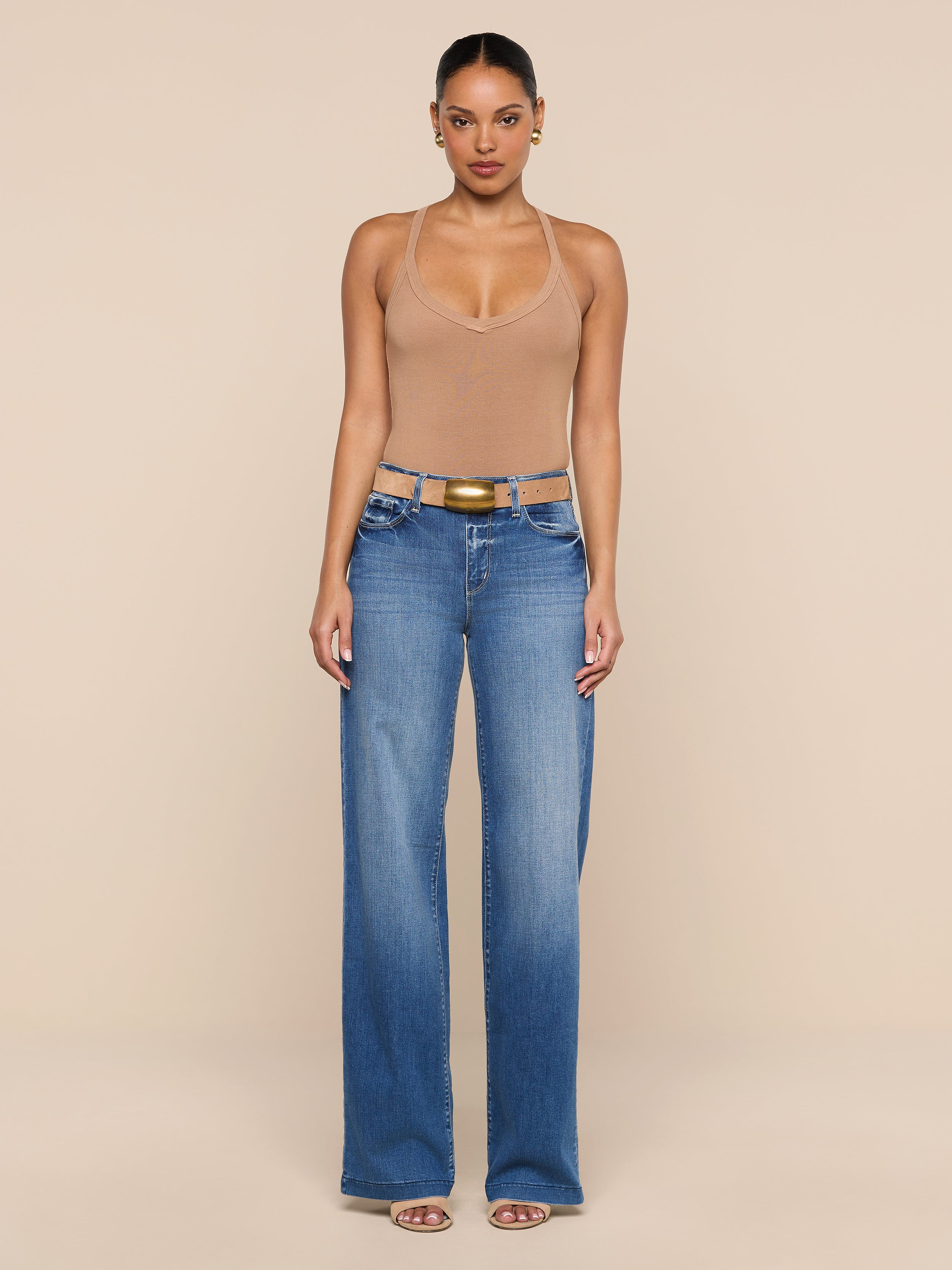 A person stands facing forward, wearing the Wrenna Racerback Bodysuit in tan, paired with blue wide-leg jeans, a brown belt, and sandals against a plain white background.