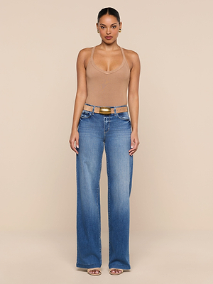 A woman faces forward in a fitted tan Wrenna Racerback Bodysuit, wide-leg blue jeans with hip cutouts, a wide belt, and heeled sandals against a plain beige background.