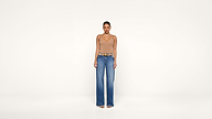 A woman stands facing forward against a plain white background, wearing a tan sleeveless top, blue wide-leg jeans, and sandals.
