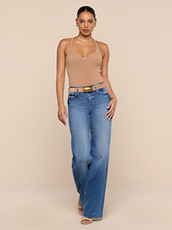 A person stands against a plain white background, wearing the Wrenna Racerback Bodysuit in beige, blue high-waisted wide-leg jeans, a belt, and sandals.