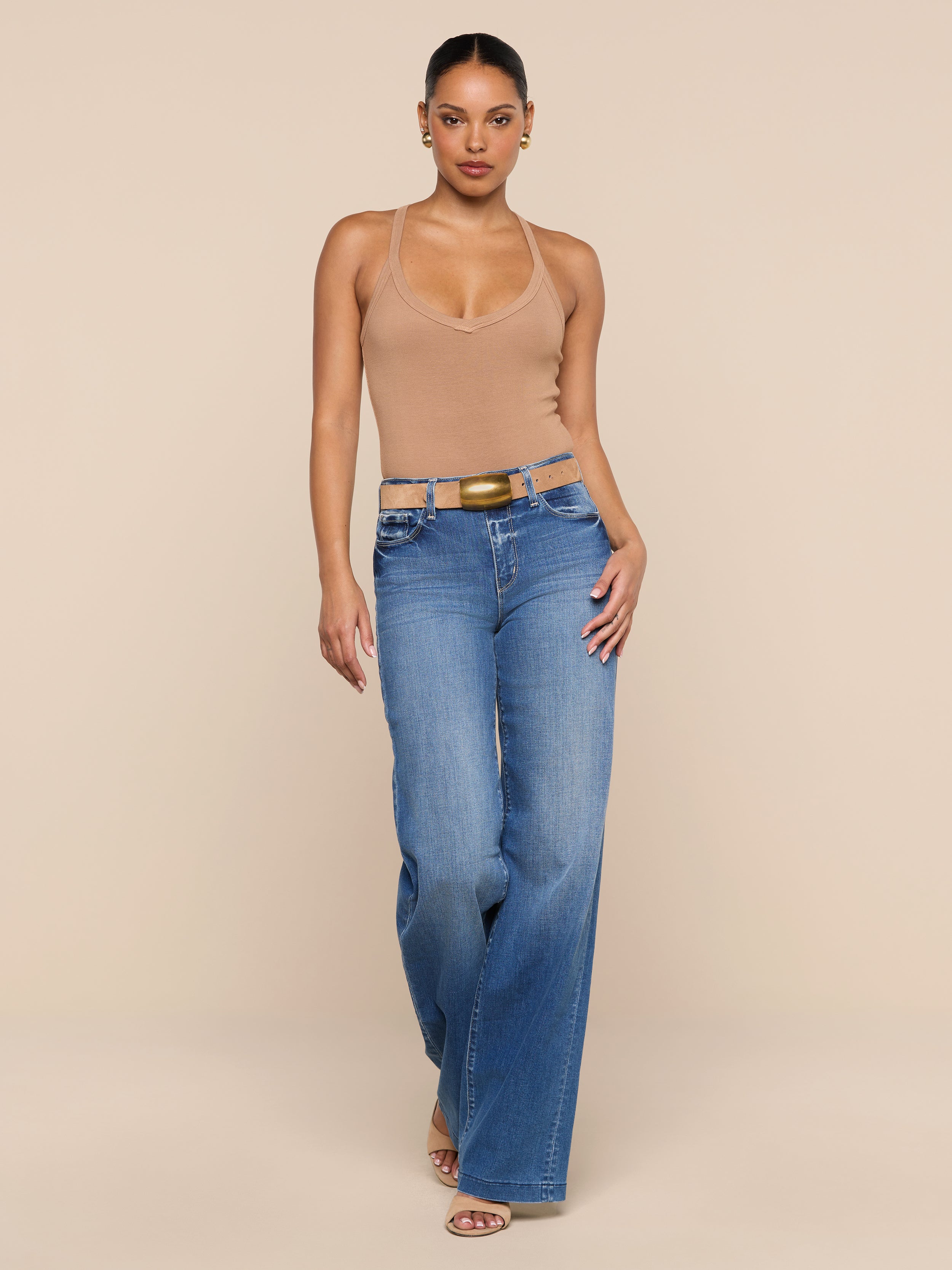 A person stands against a plain white background, wearing the Wrenna Racerback Bodysuit in beige, blue high-waisted wide-leg jeans, a belt, and sandals.