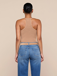 A person with their hair in a bun, wearing the Wrenna Racerback Bodysuit in tan rib fabric and blue jeans, stands facing away from the camera against a plain, light background.